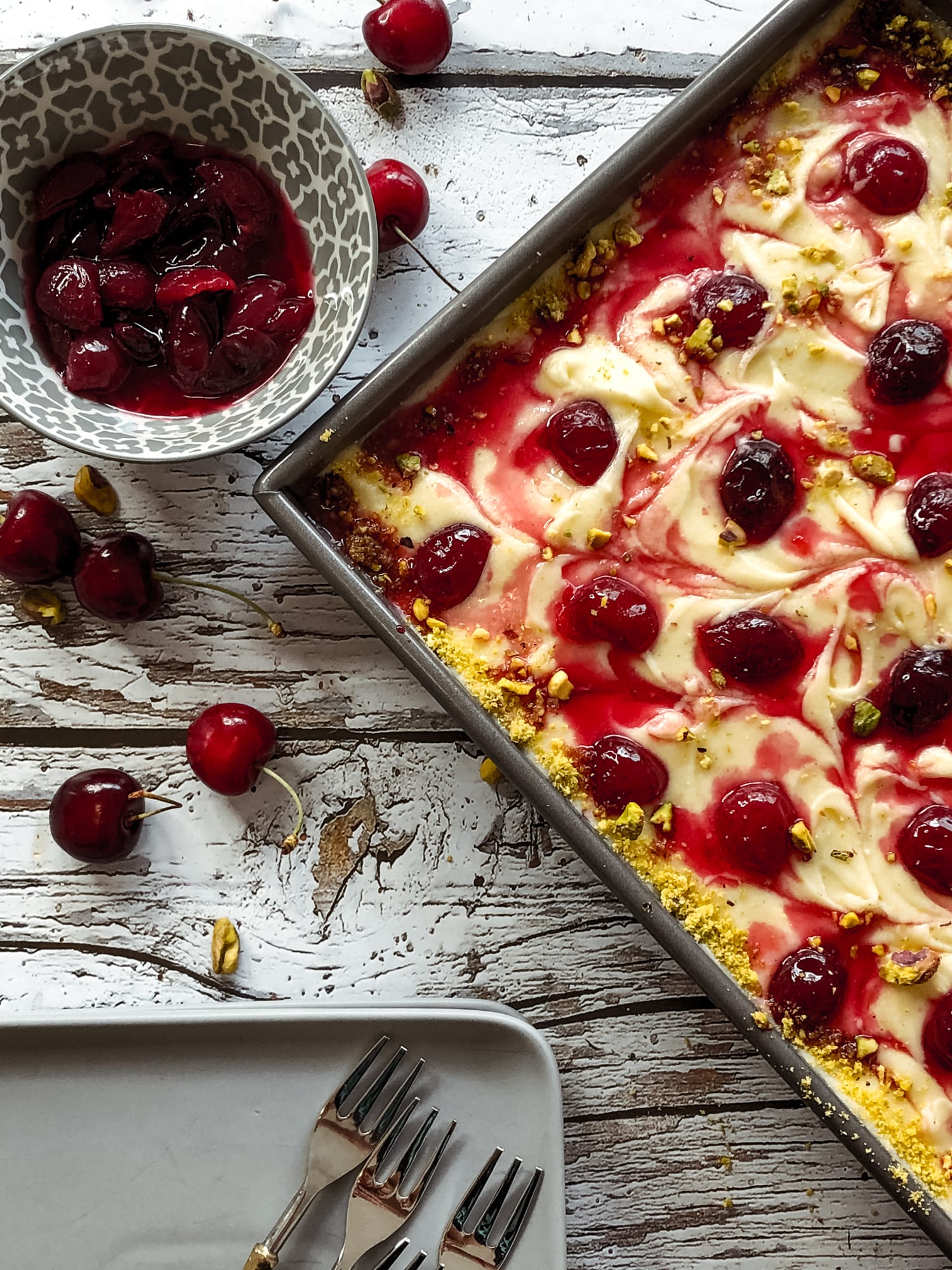 A corner of a pistachio and cherry sheet cake beside a bowl of cherries and a plate of forks