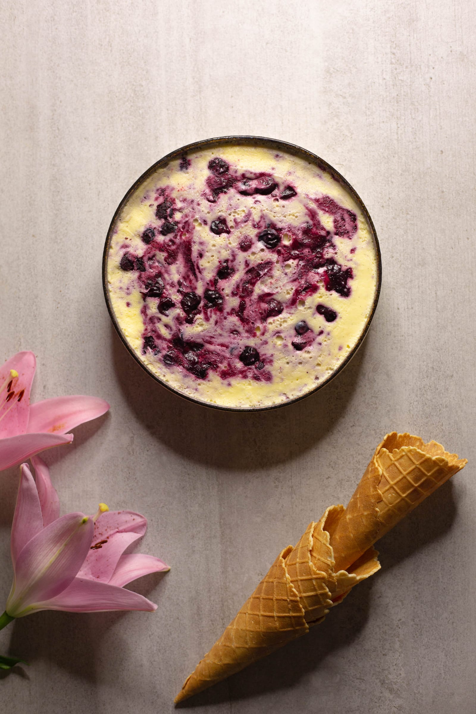 A tin of blueberry swirl ice cream before scooping, with a waffle cone and lilies, from above