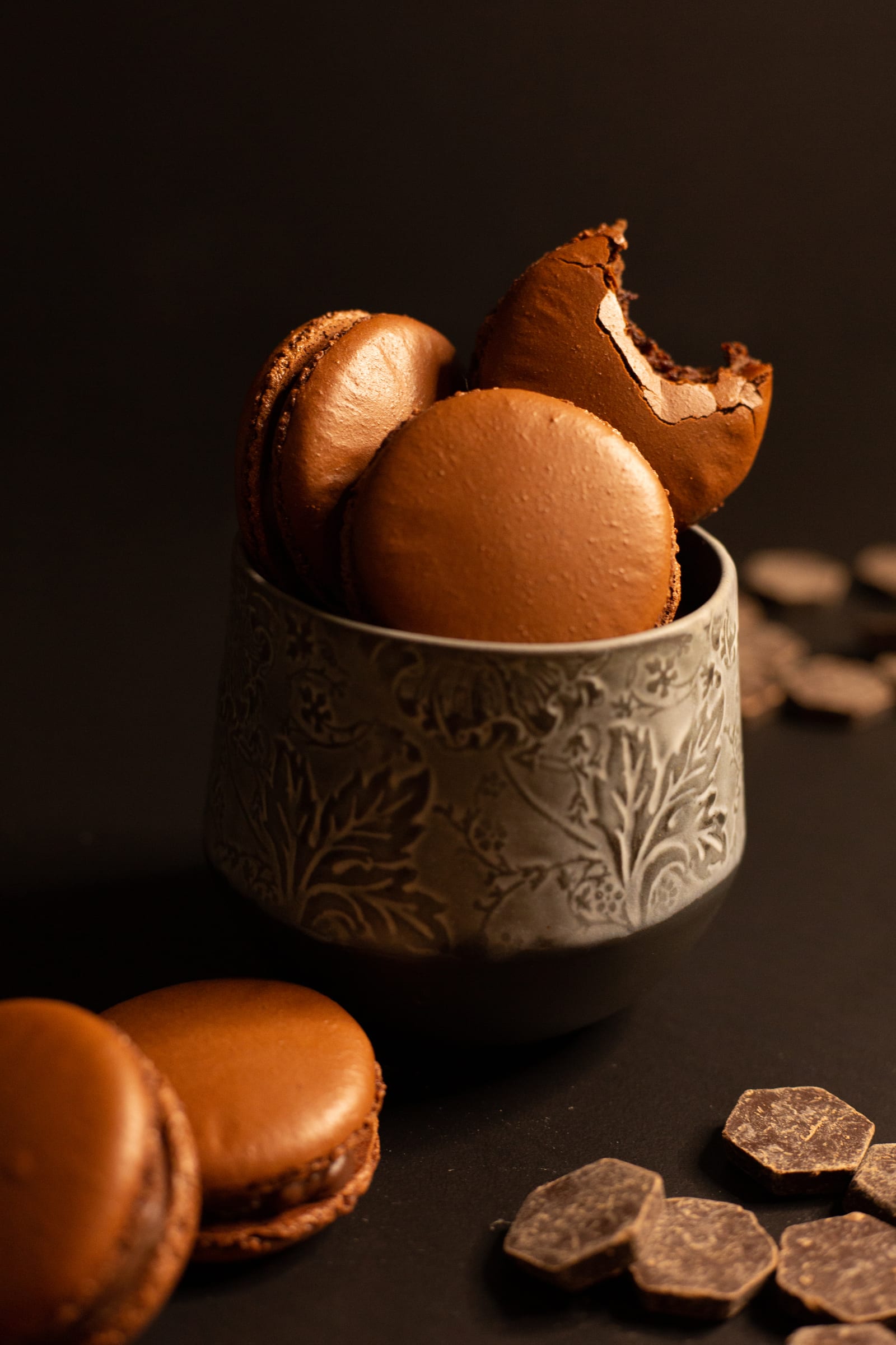 Chocolate macarons stacked in an embossed cup, the top one bitten to show the ganache