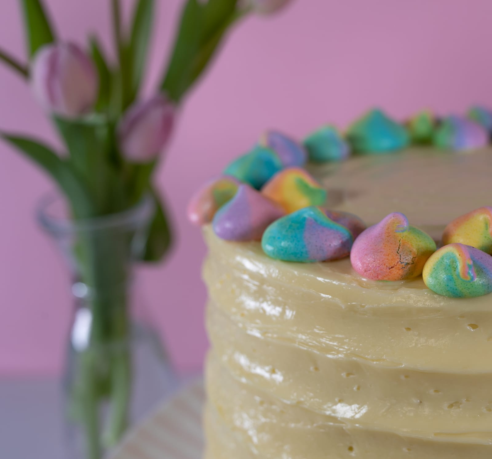 Close-up of the meringue kisses on the edge of a pastel rainbow cake, pink tulips behind