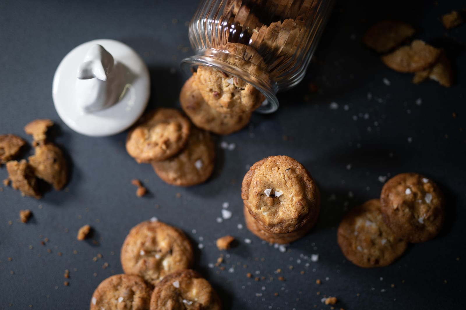 Pecan and white chocolate salted cookies spilling from a tipped jar beside its bunny lid, from above