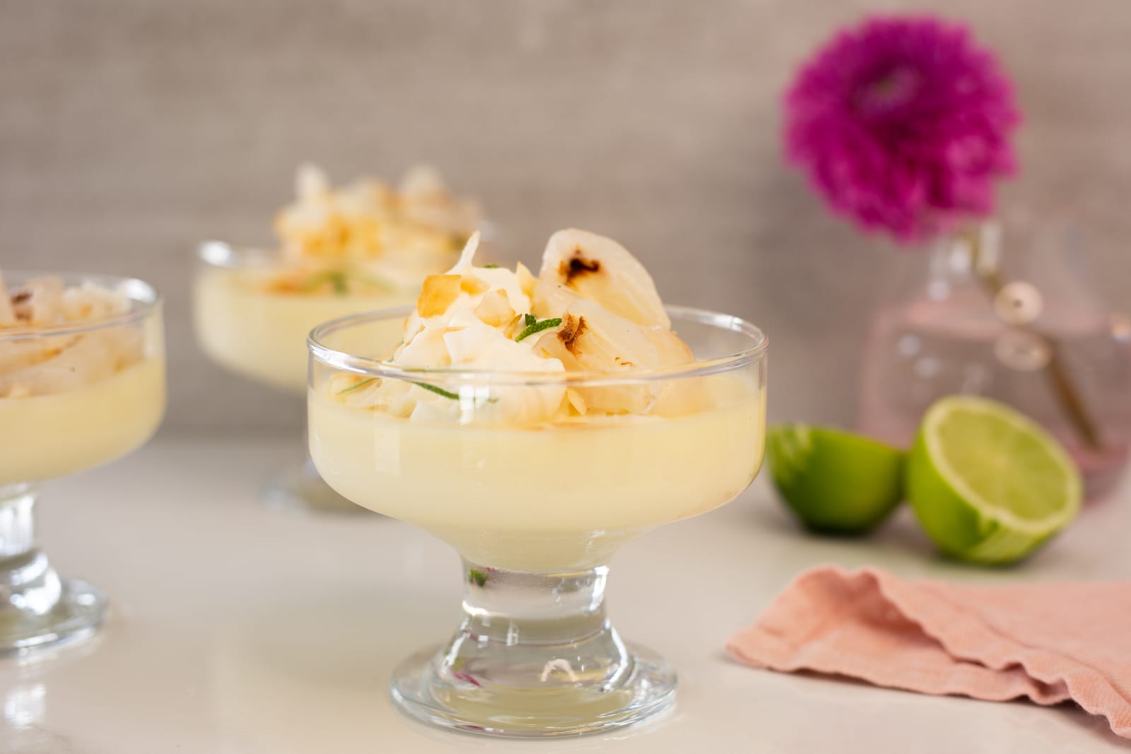 Lime and coconut possets with litchis in footed glasses beside halved limes and a pink dahlia