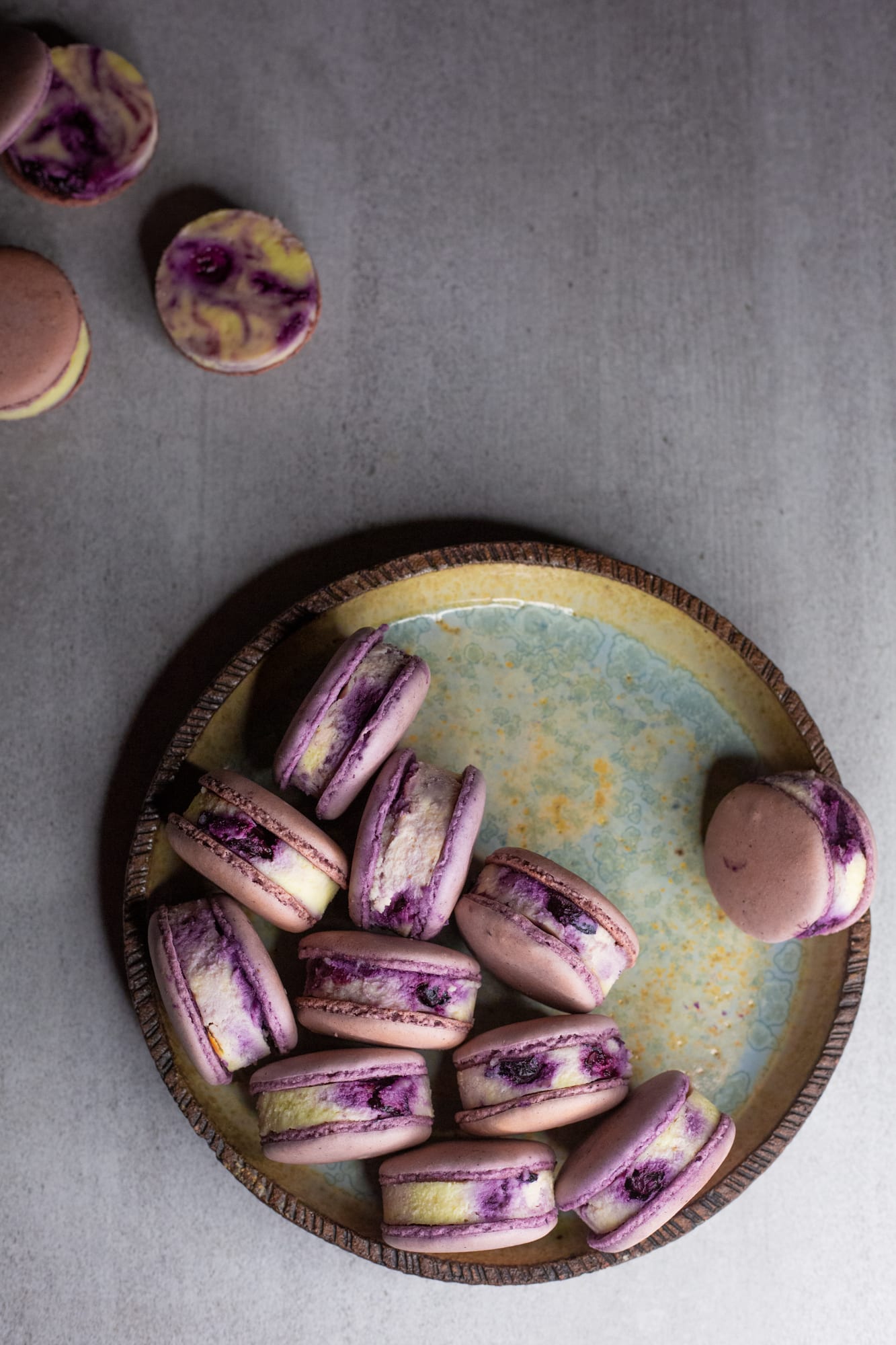 A speckled plate of blueberry cheesecake macarons with more scattered beside it, from above
