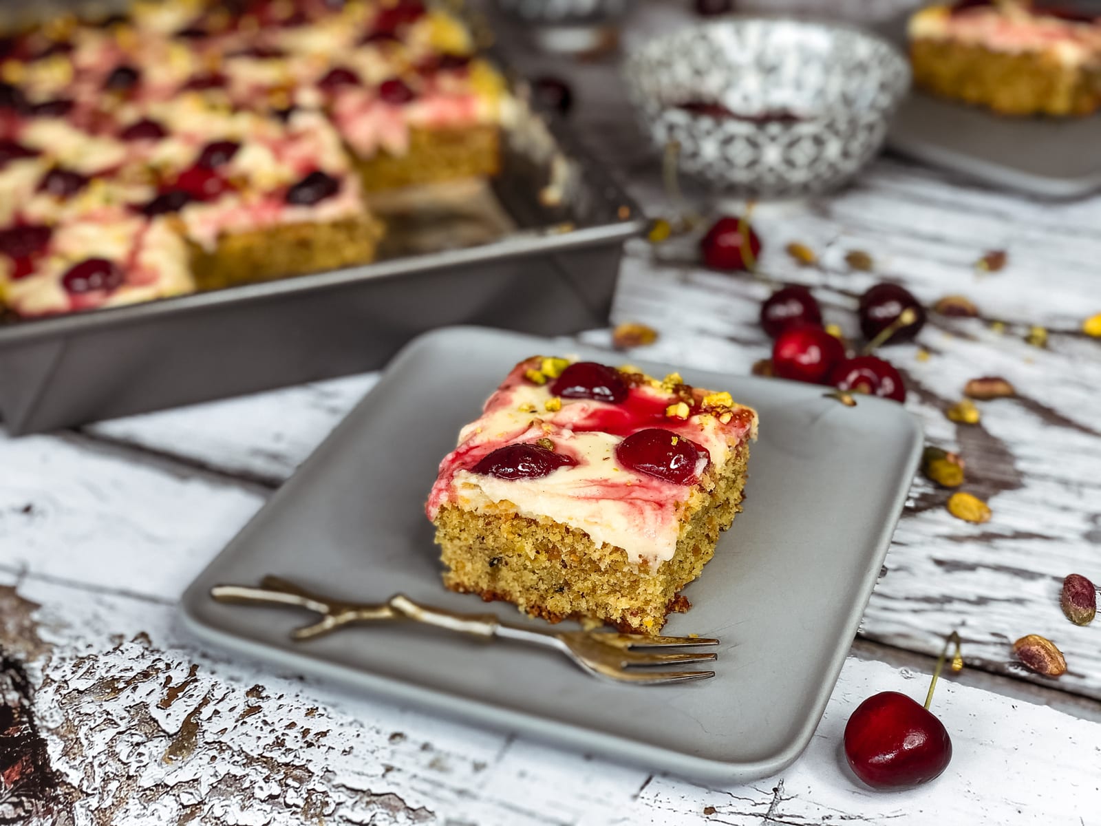 A square of pistachio and cherry sheet cake on a grey plate with a gold fork, the cut tin behind