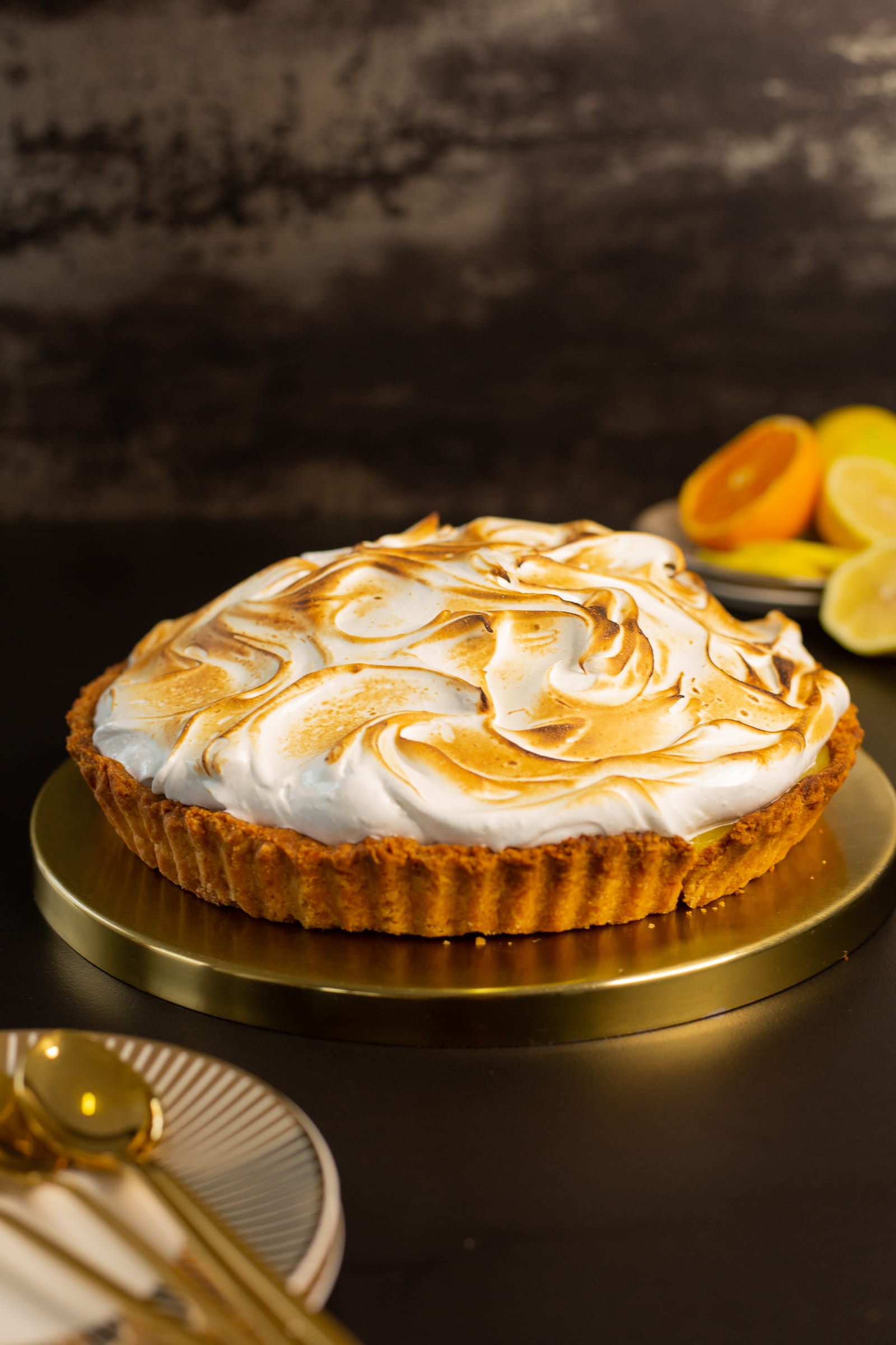 A whole lemon meringue pie on a gold board with lemons and oranges behind