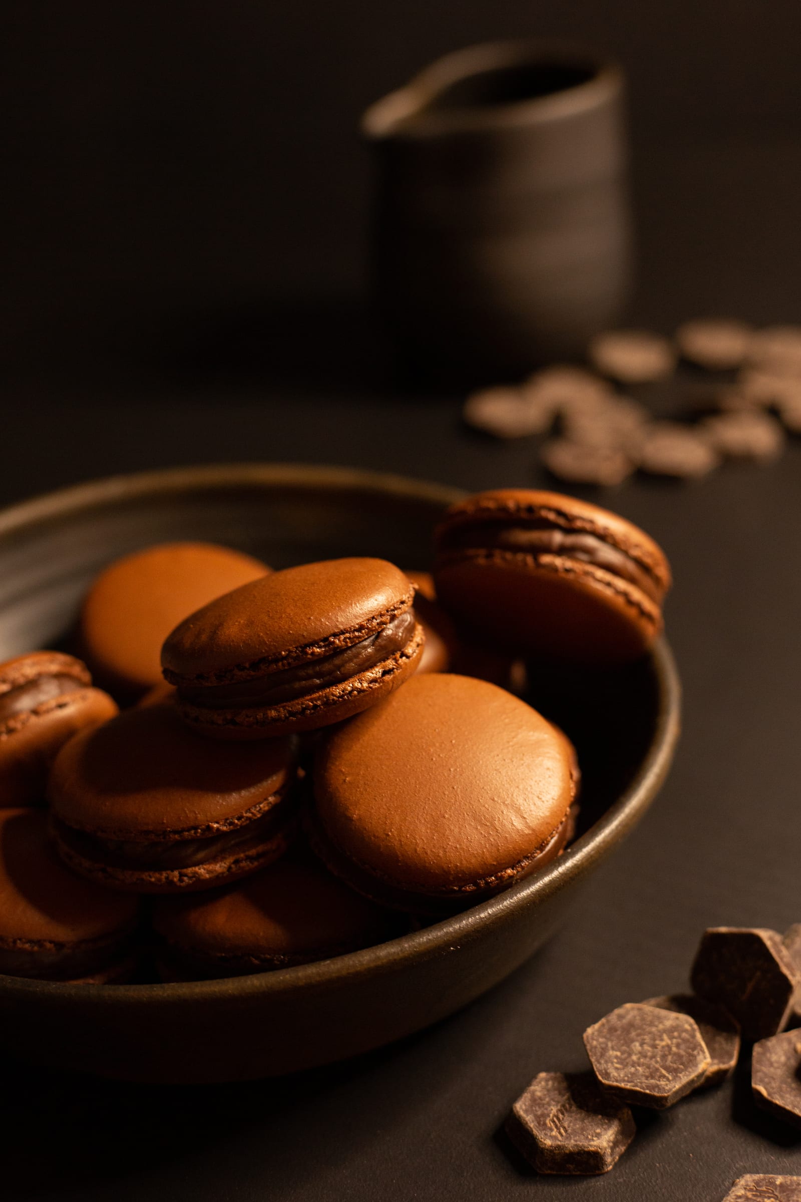 A dark bowl of chocolate macarons with chocolate pieces scattered around it