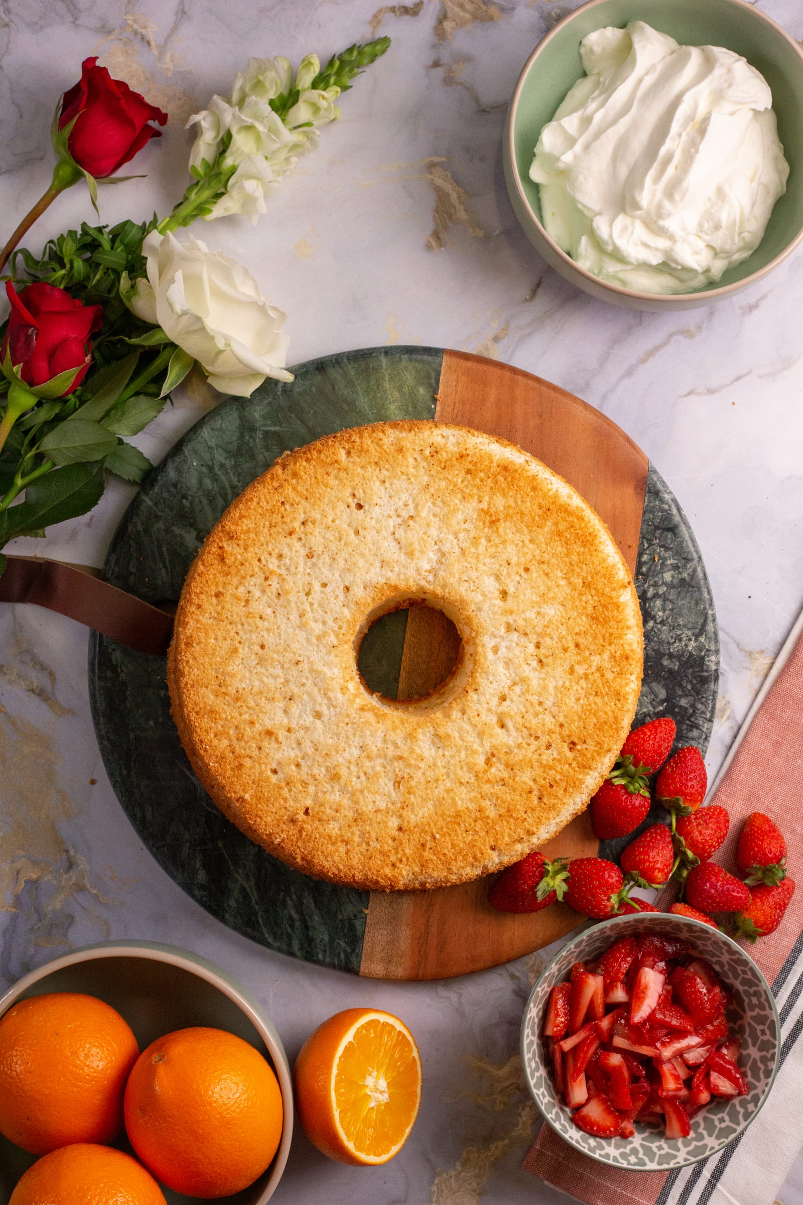 The baked angel food cake ring on a marble board, bowls of cream and strawberries around it, from above