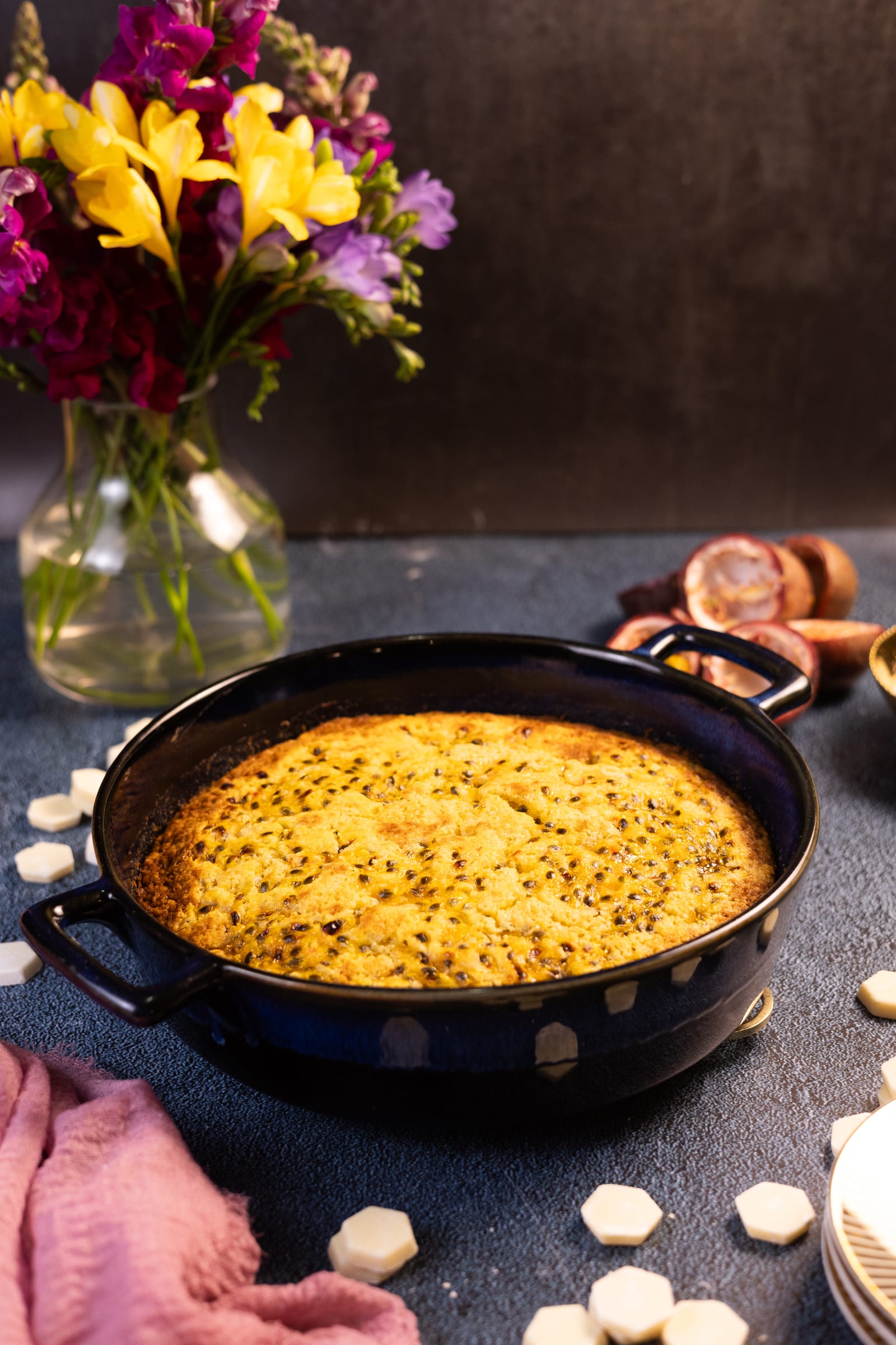 The baked passionfruit pudding in a dark pot, with flowers and passionfruit