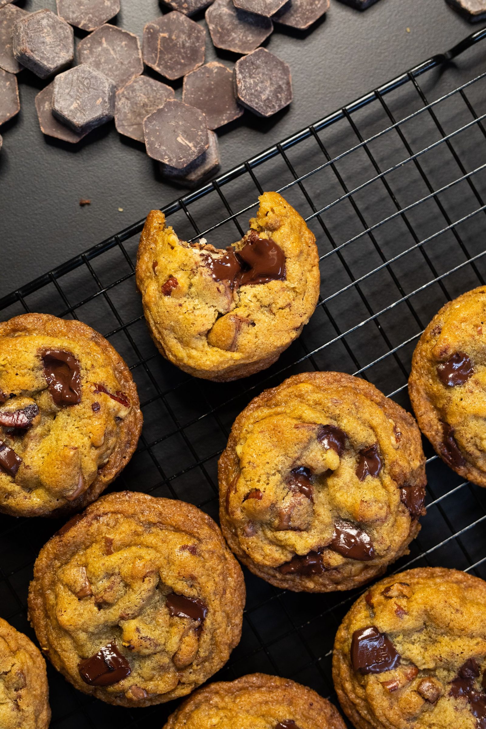 A bitten chocolate chip cookie with a soft centre on a cooling rack, below a pile of chocolate chunks, from above