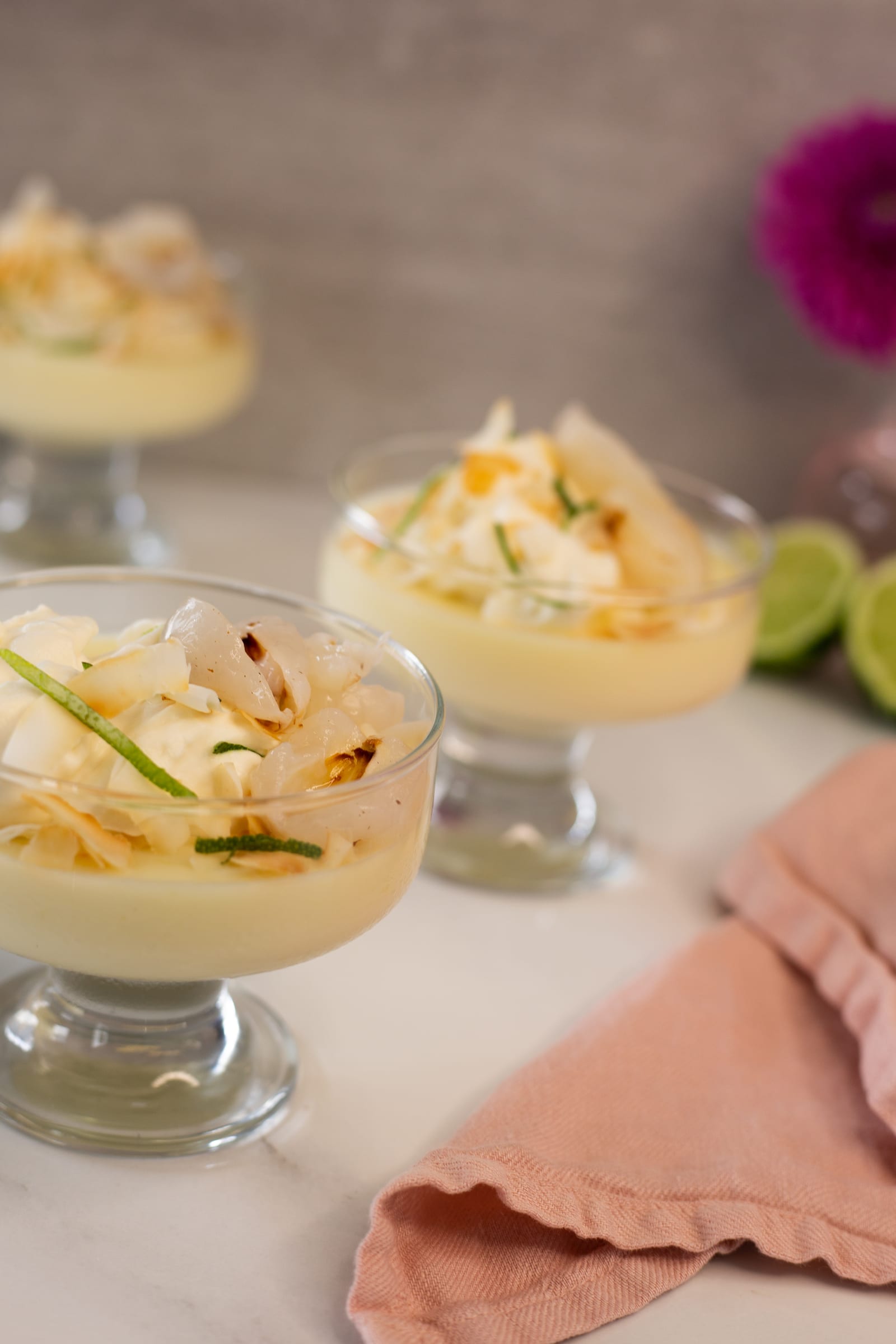 Three lime and coconut possets with litchis seen from above, with a pink napkin in front