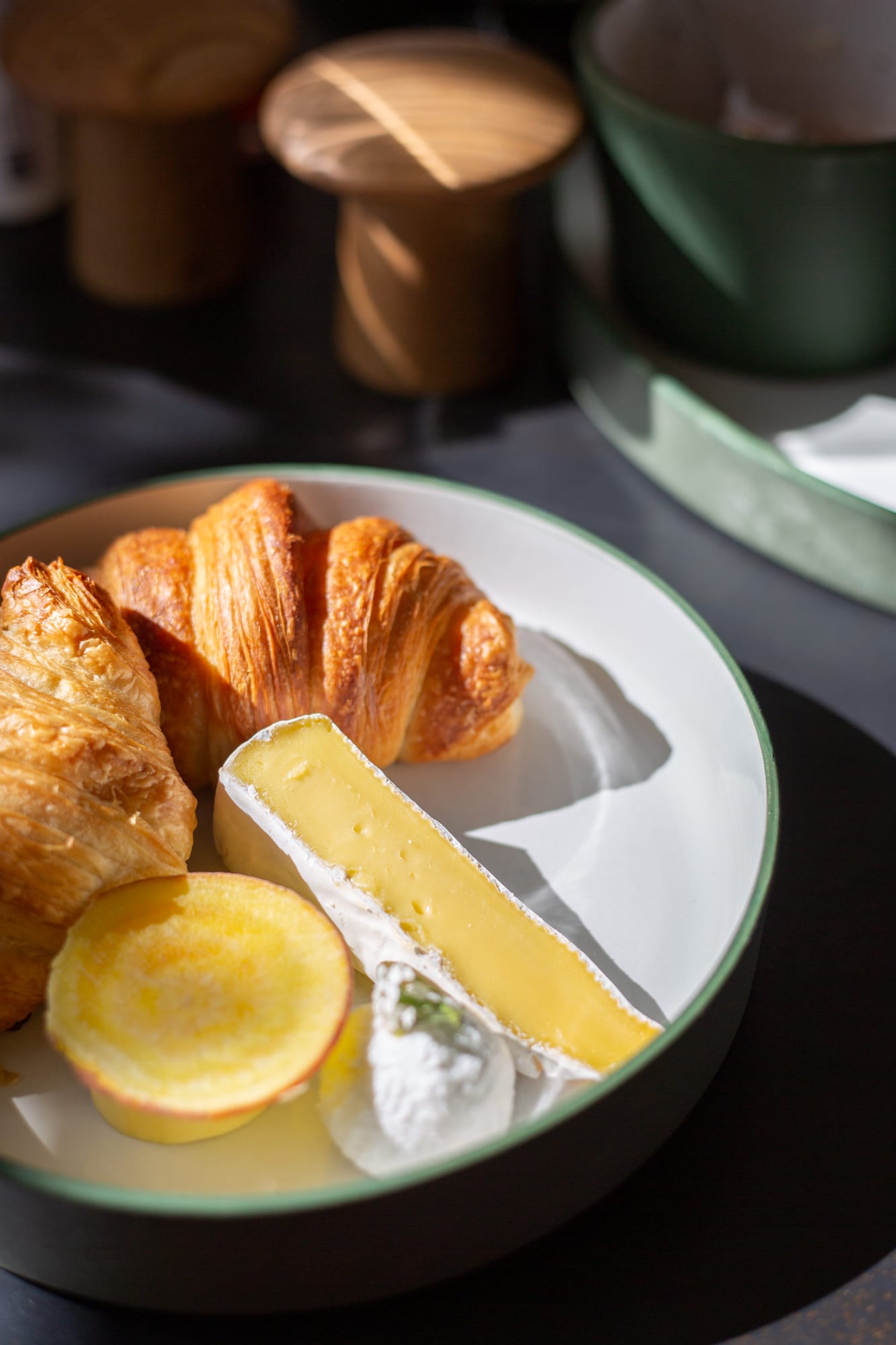 Croissants, a wedge of brie, a golden beetroot slice and a dollop of cream cheese in a green-rimmed bowl in morning sun