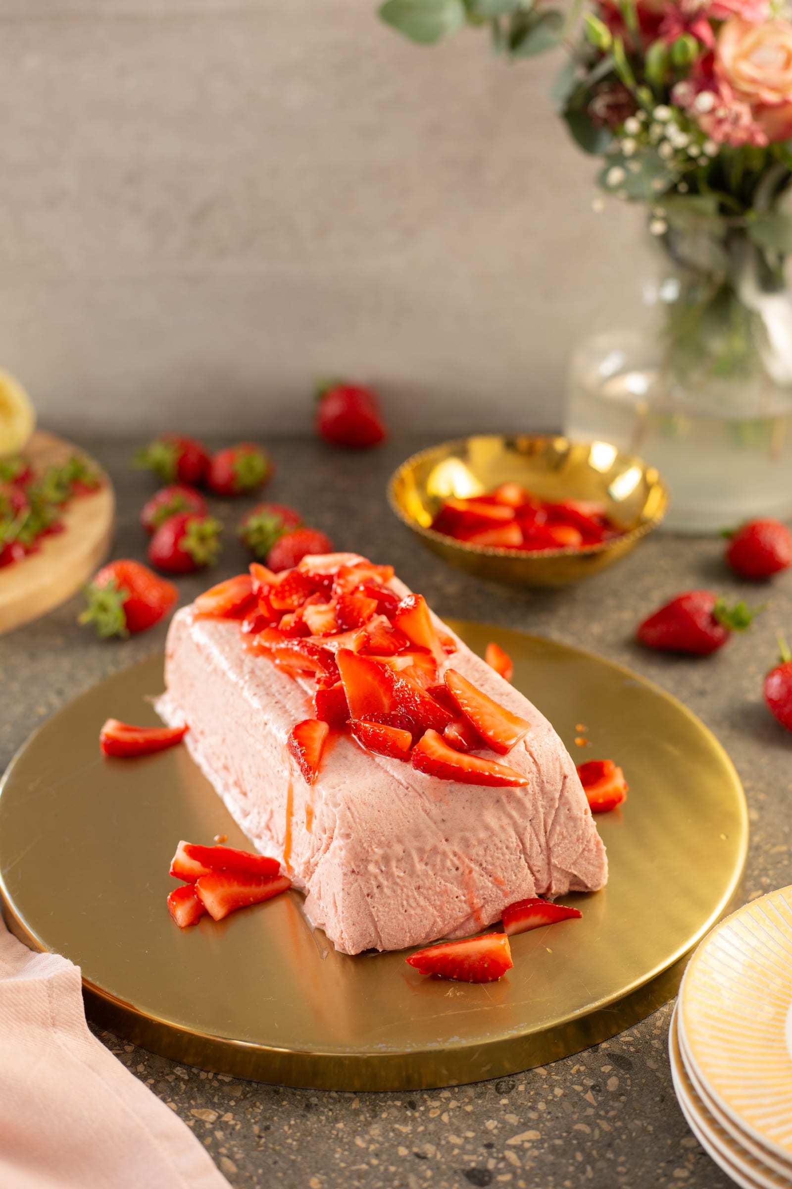 A roasted strawberry and rosemary semifreddo on a gold plate with flowers and bowls of strawberries