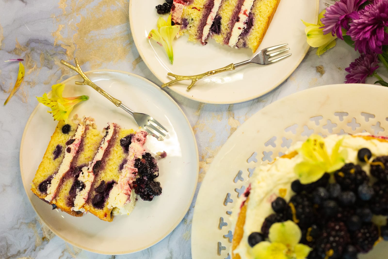 A slice of lemon and blueberry cake on a plate with a fork beside the top of the cake, from above