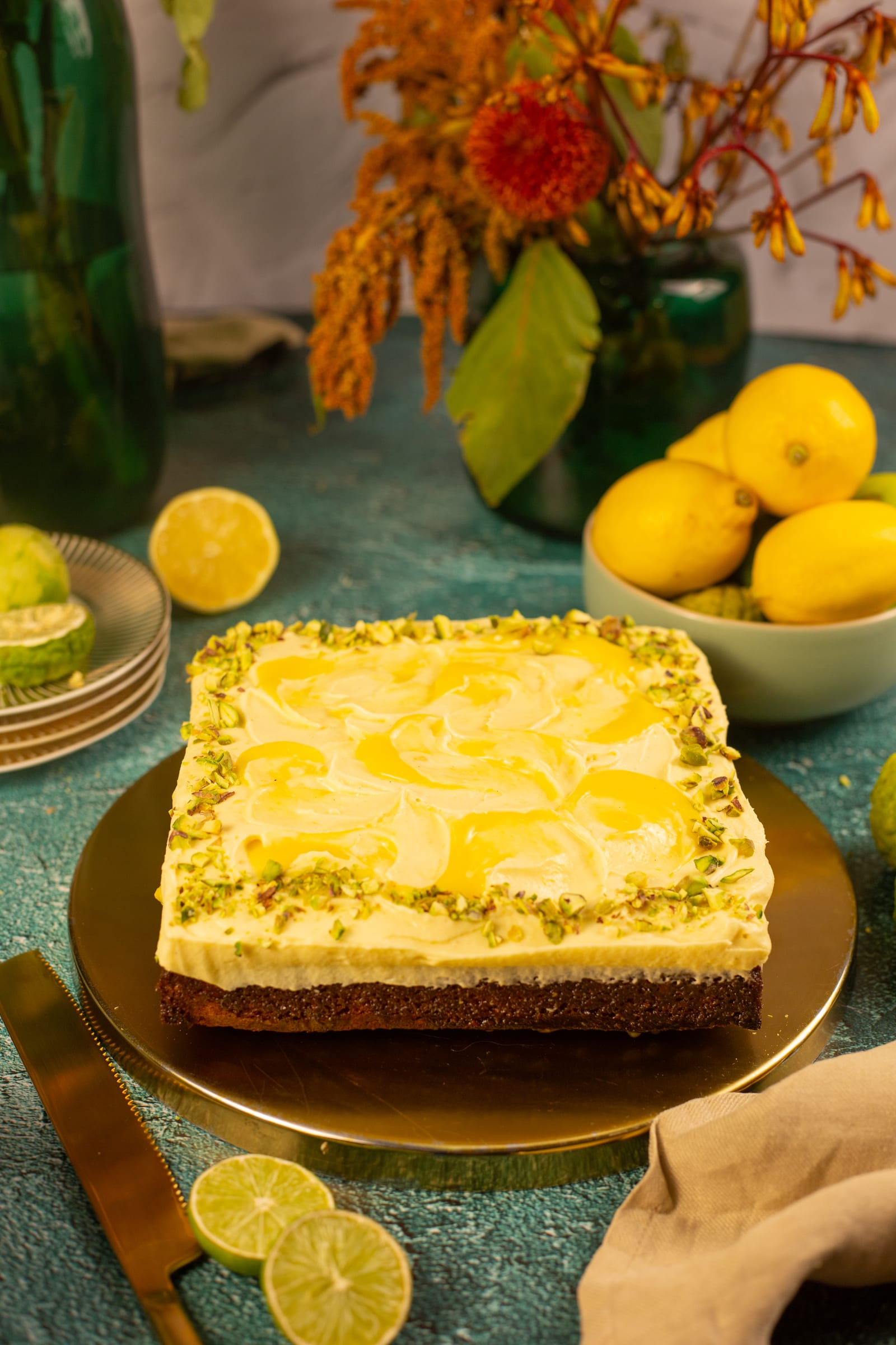 A pistachio and lemon traybake on a gold board, with orange flowers and a bowl of lemons
