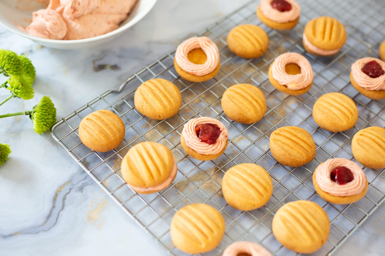 Custard and jelly biscuits, some sandwiched with jam rings, on a wire rack beside a bowl of filling, from above