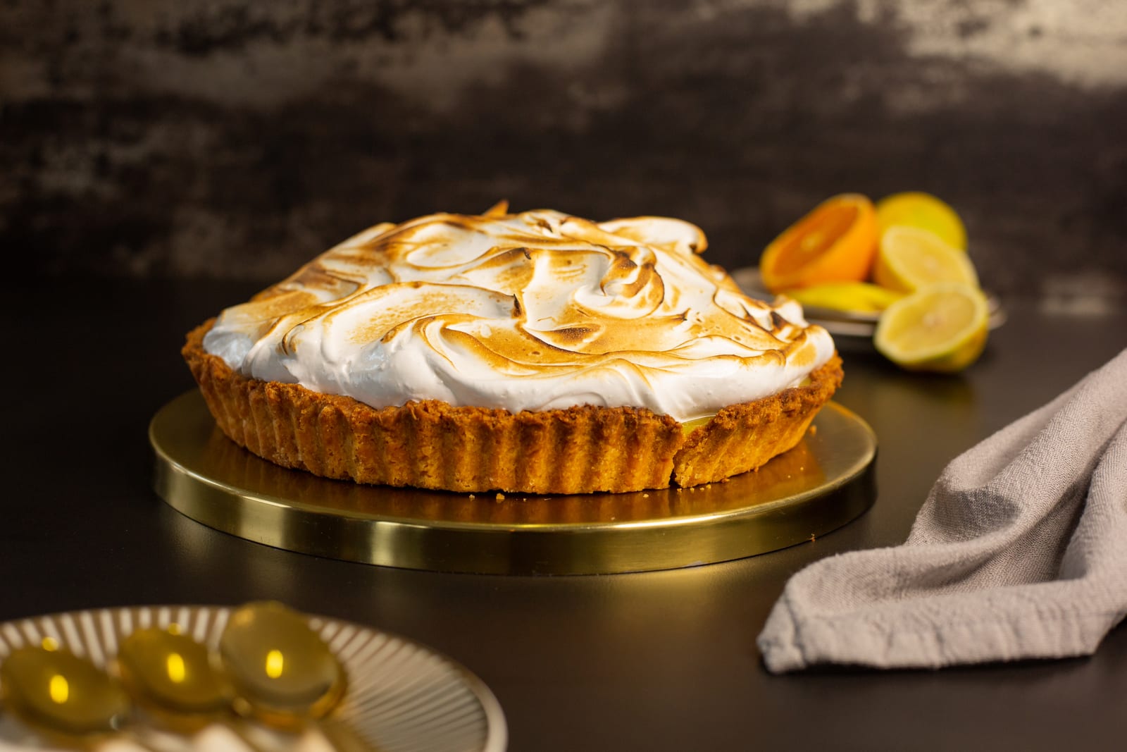 A whole lemon meringue pie with toasted meringue swirls, with citrus and a grey napkin beside it