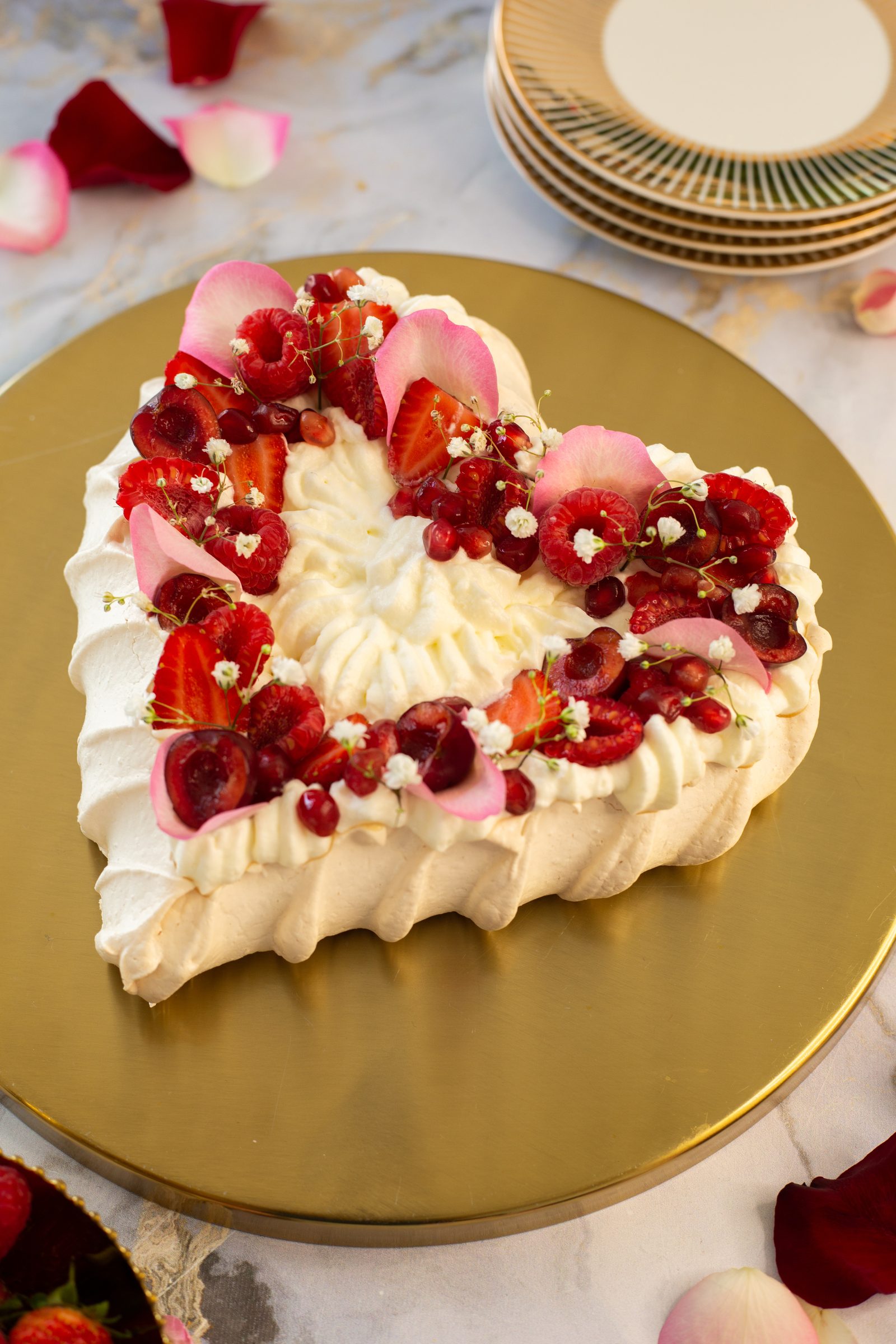 A heart-shaped berry pavlova on a gold cake board with stacked plates and rose petals, from a high angle