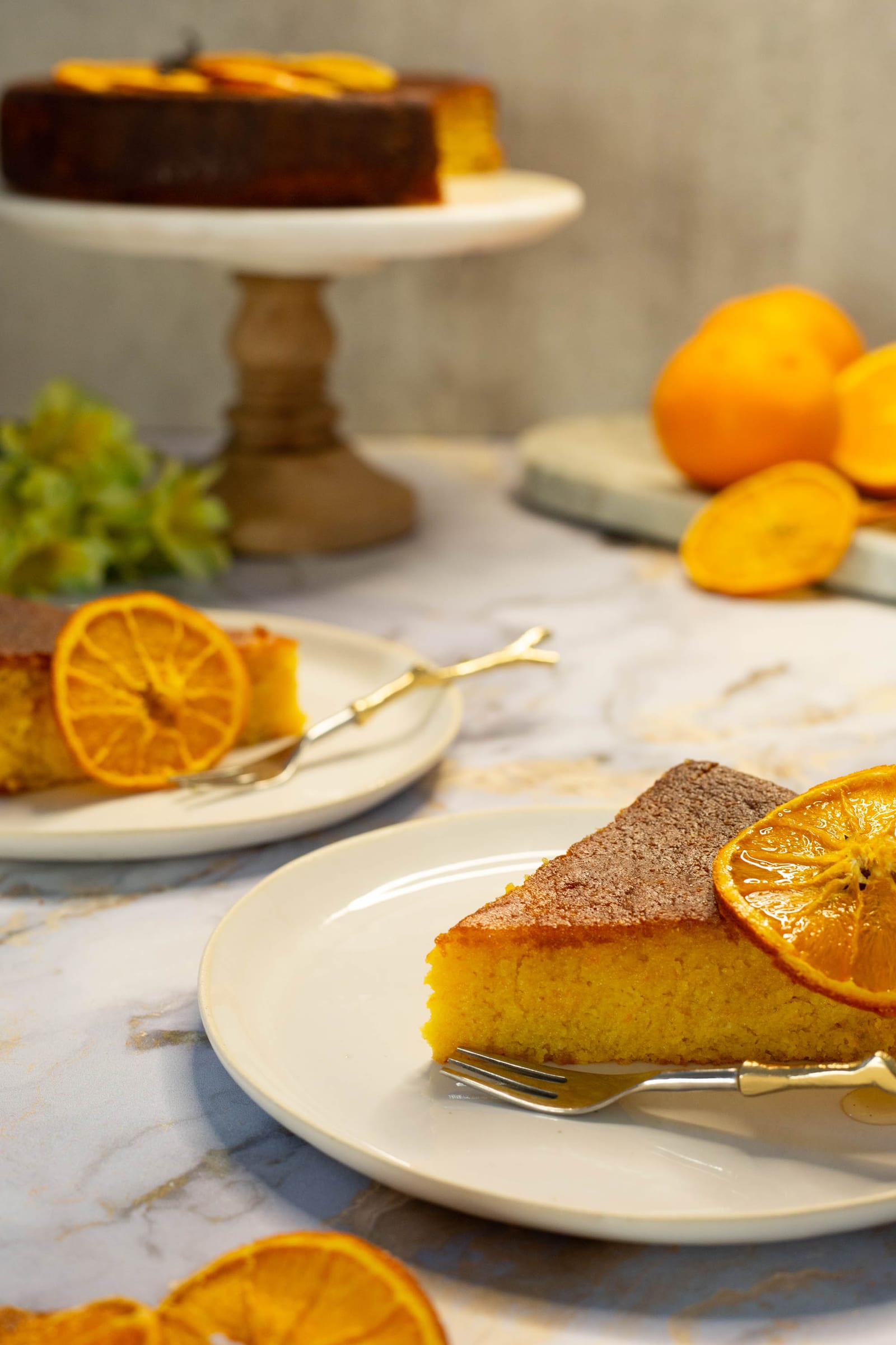 Two slices of whole orange, cardamom and polenta cake on plates, the cut cake on its stand behind