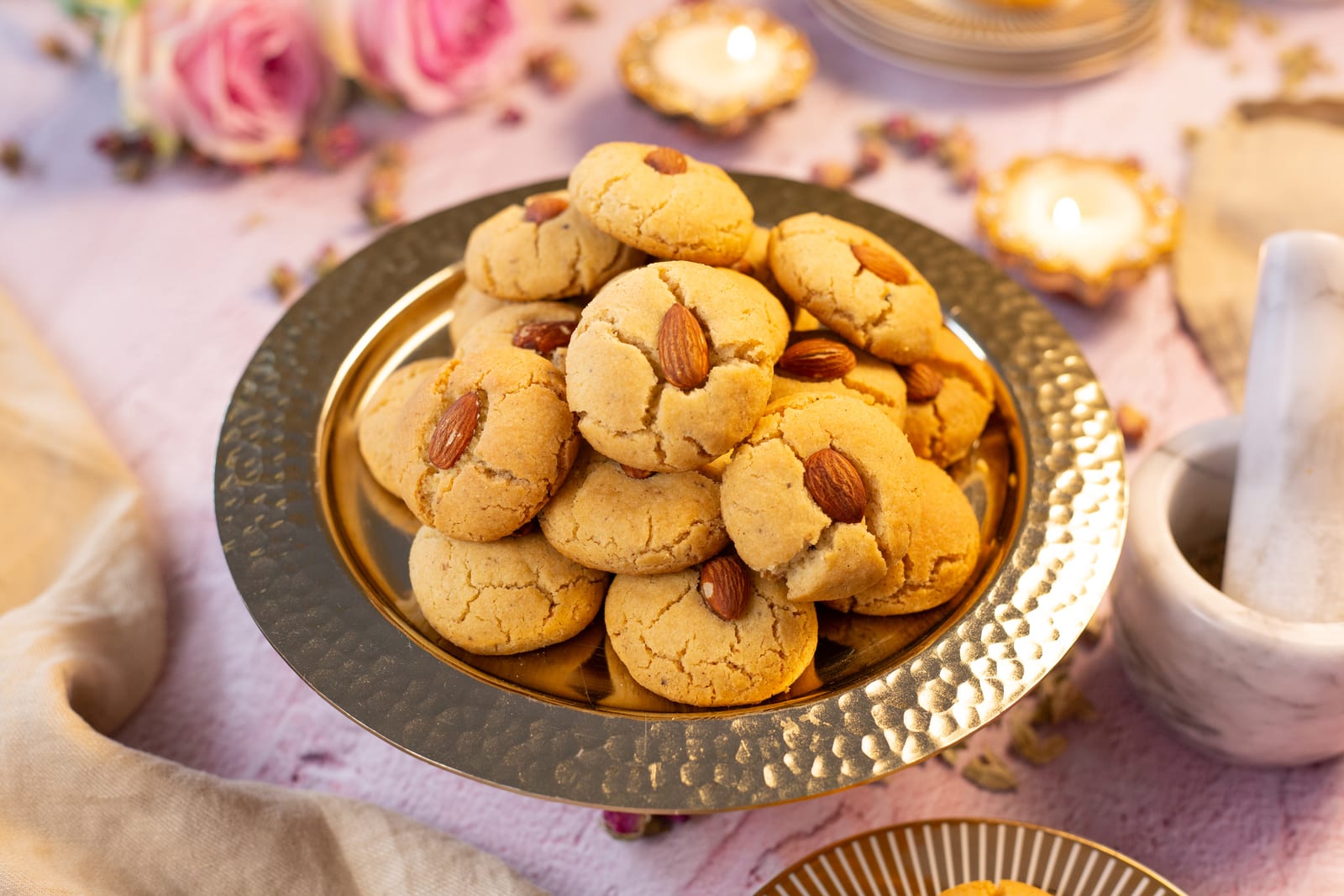 Naan kathai biscuits topped with almonds in a hammered brass bowl, with roses and diyas