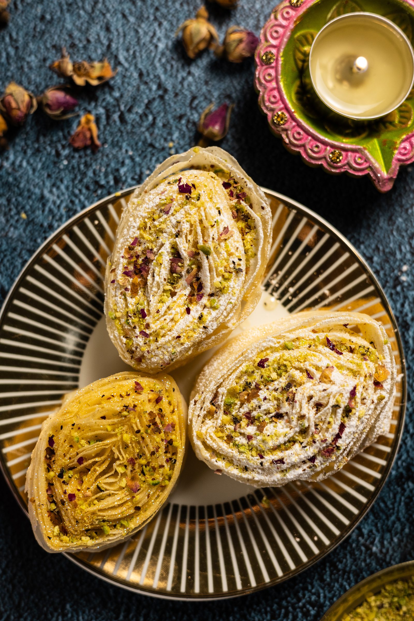 Three banana puri on a striped gold plate with a diya, rosebuds and cardamom, from above