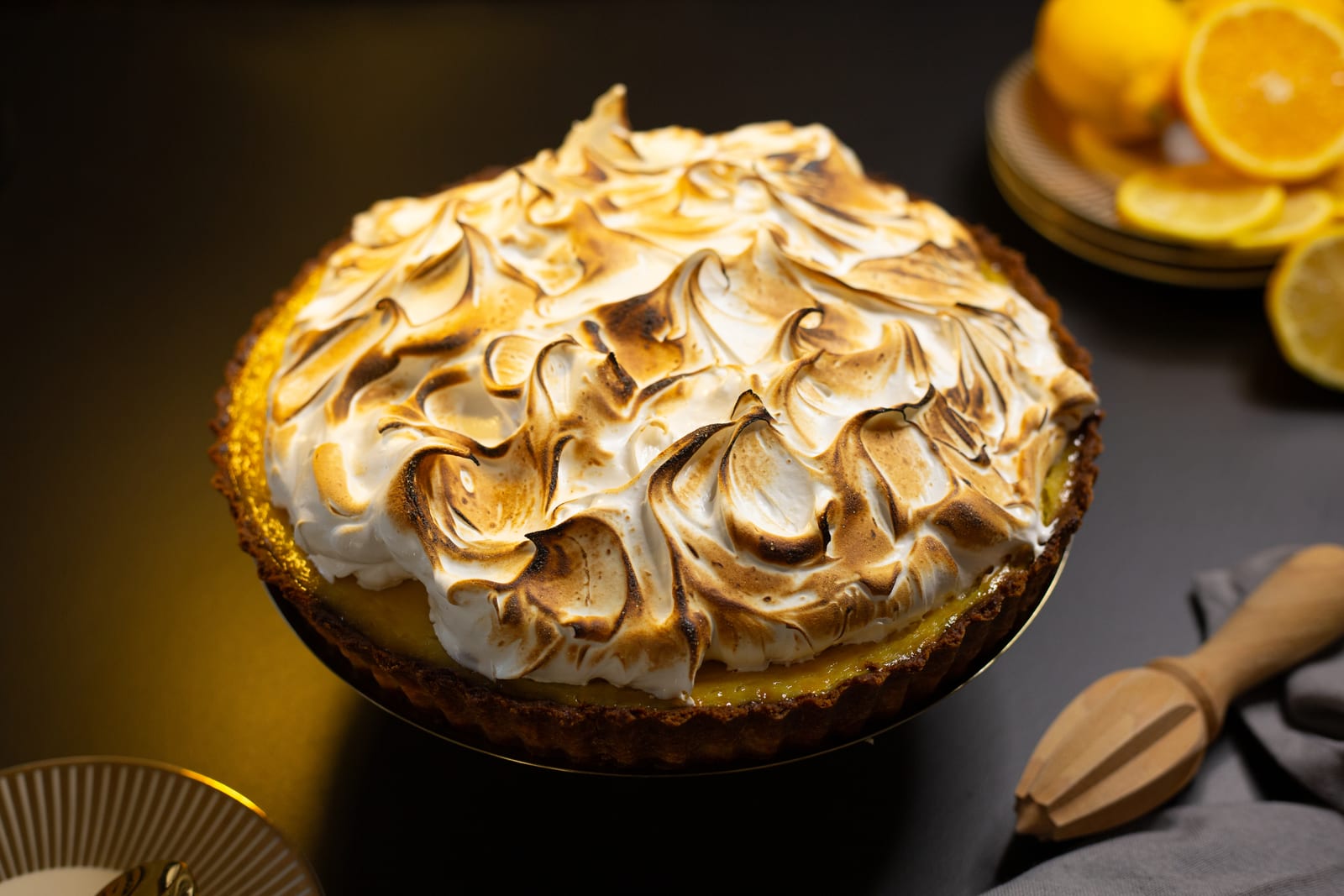 A lemon meringue pie piled with toasted Italian meringue, with lemons on a dark table