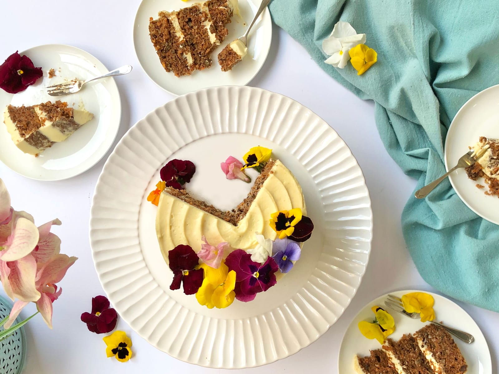 A cut carrot cake with a wedge taken out and slices on side plates with forks, pink flowers around, from above