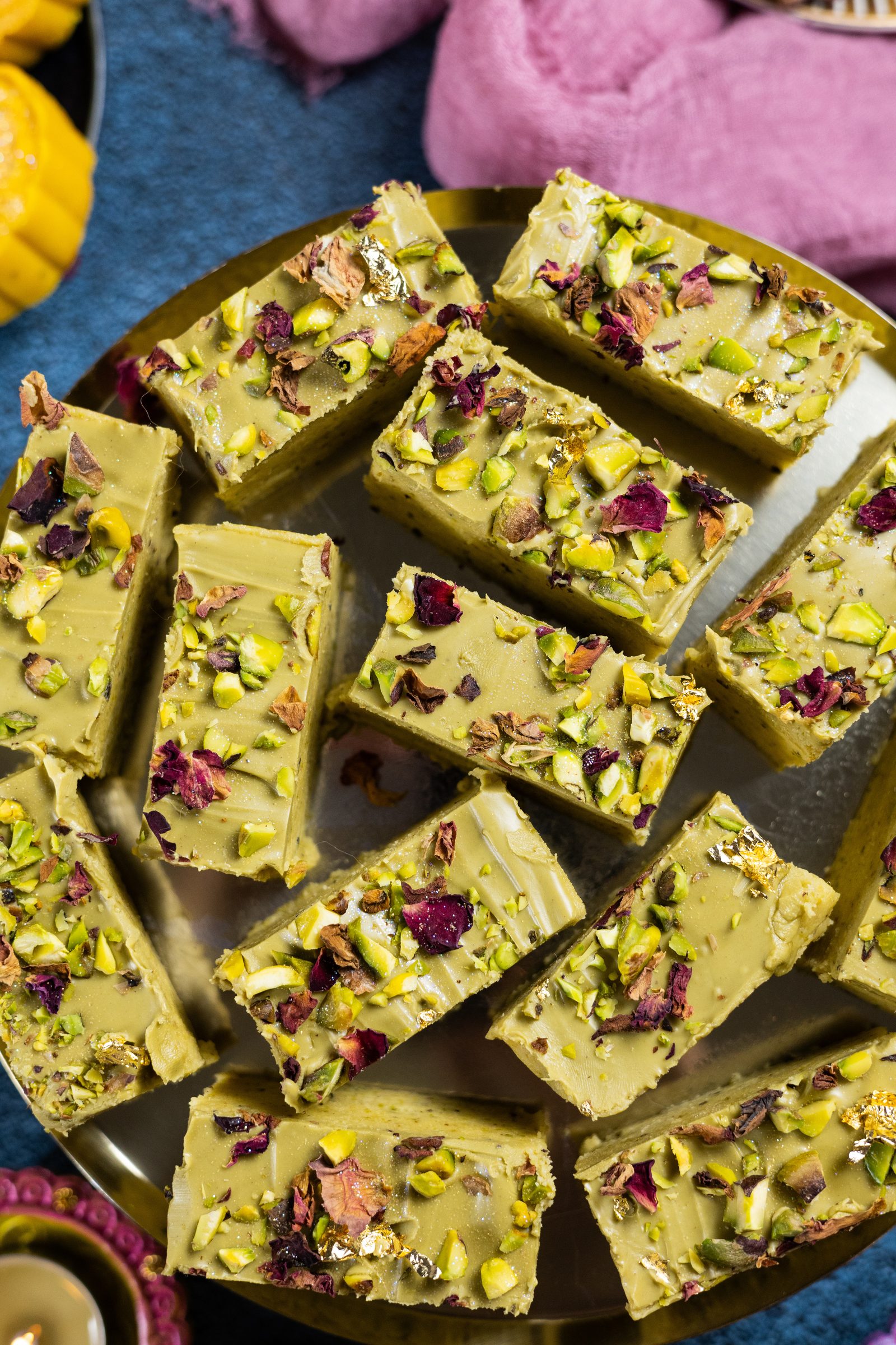 Pistachio burfee squares on a gold plate with rose petals and chopped pistachios, from above