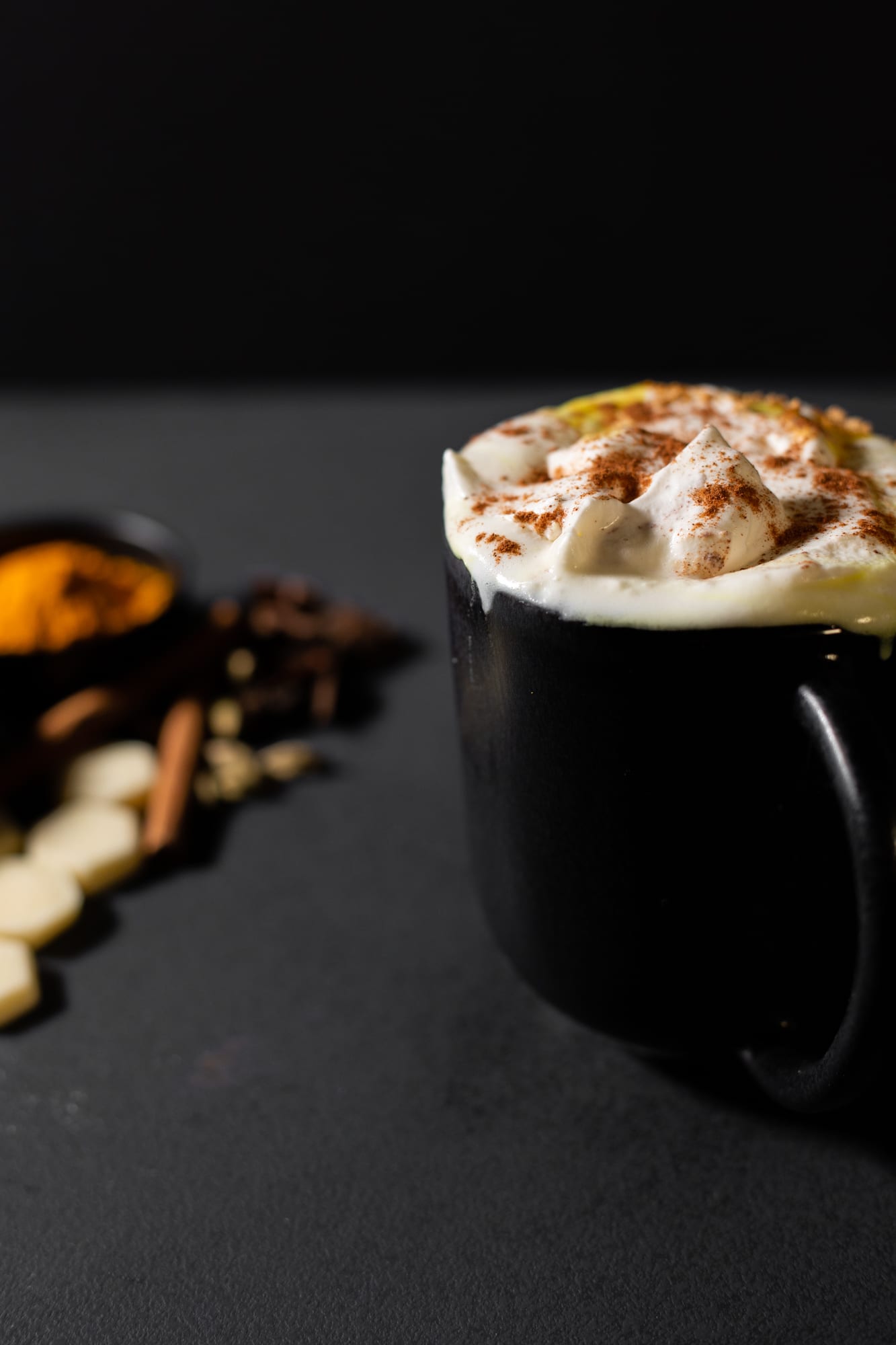 A cream-topped mug of turmeric and ginger white hot chocolate with spices beside it
