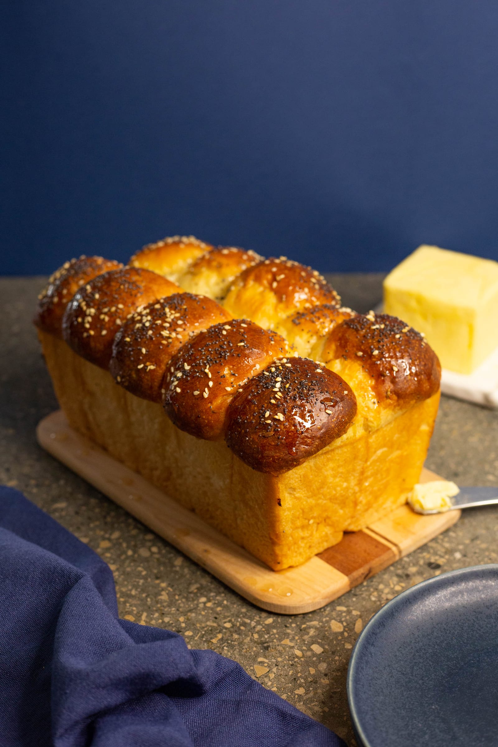 A golden mosbolletjies loaf on a wooden board with butter and blue plates