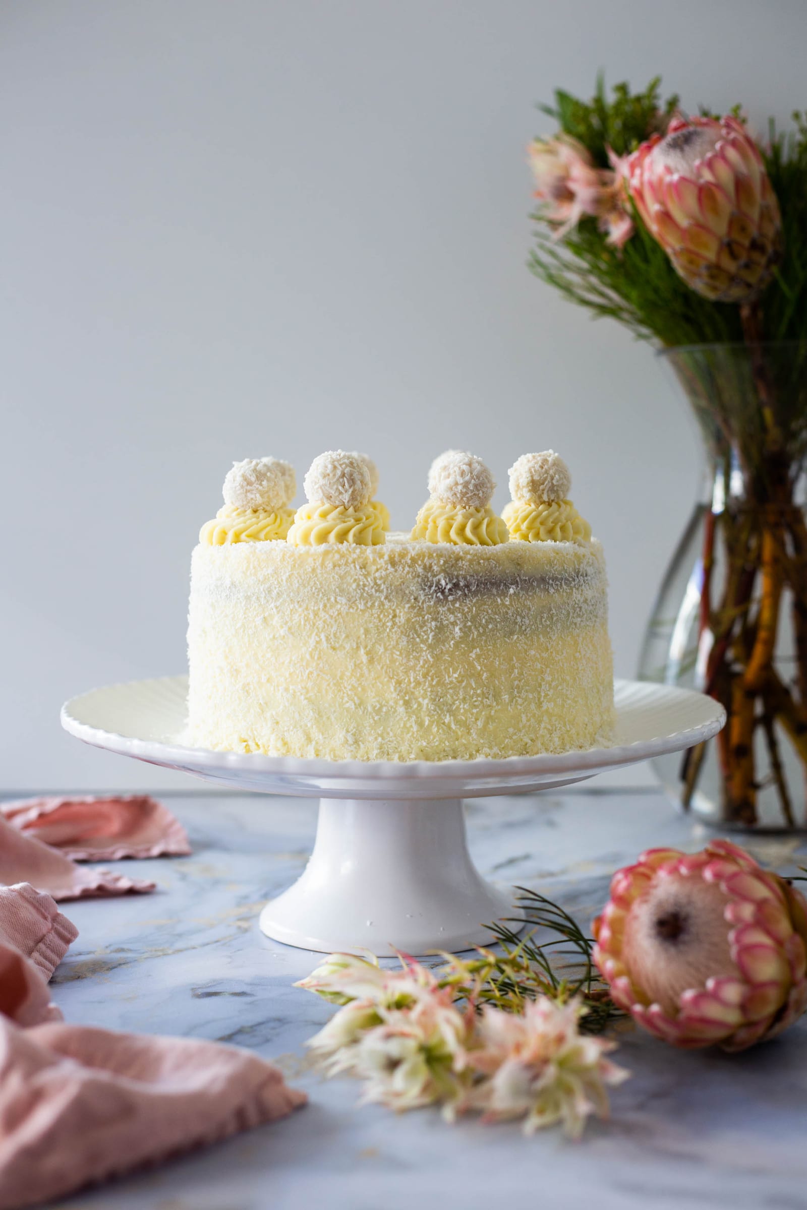 A coconut-covered Raffaello cake on a white cake stand with proteas and a pink cloth