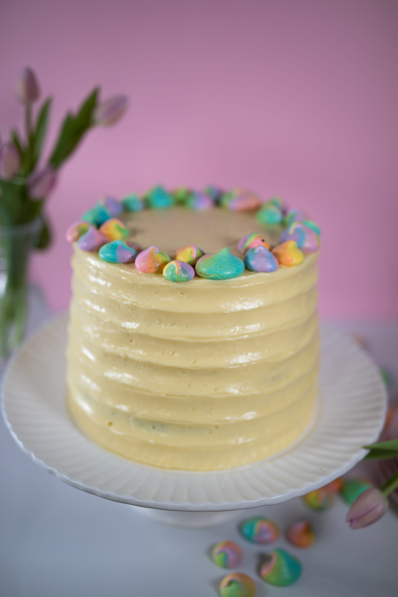 A whole pastel rainbow cake with ridged pale yellow frosting and a ring of meringue kisses, pink tulips behind