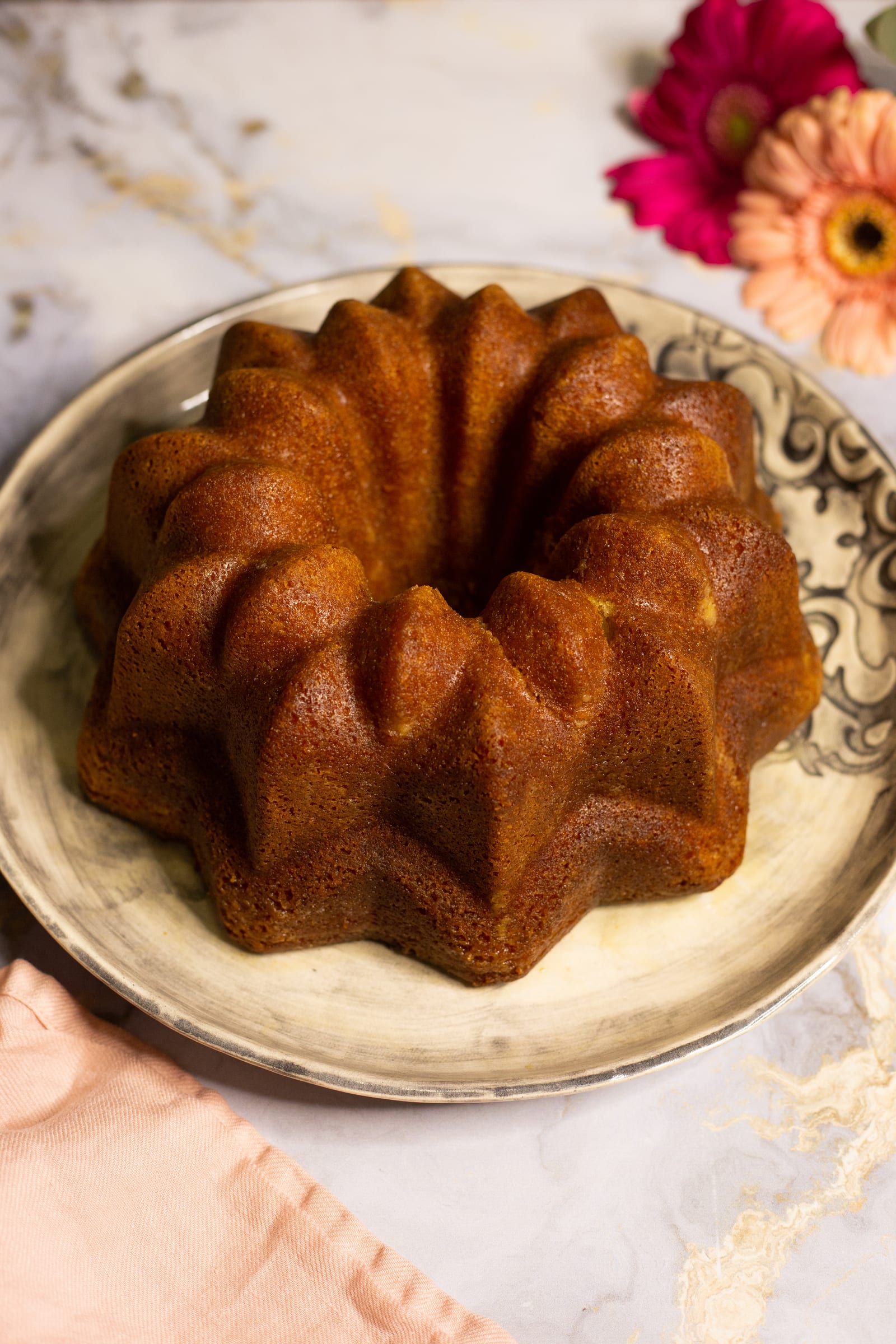 A whole Kentucky butter cake on a patterned plate beside pink gerberas