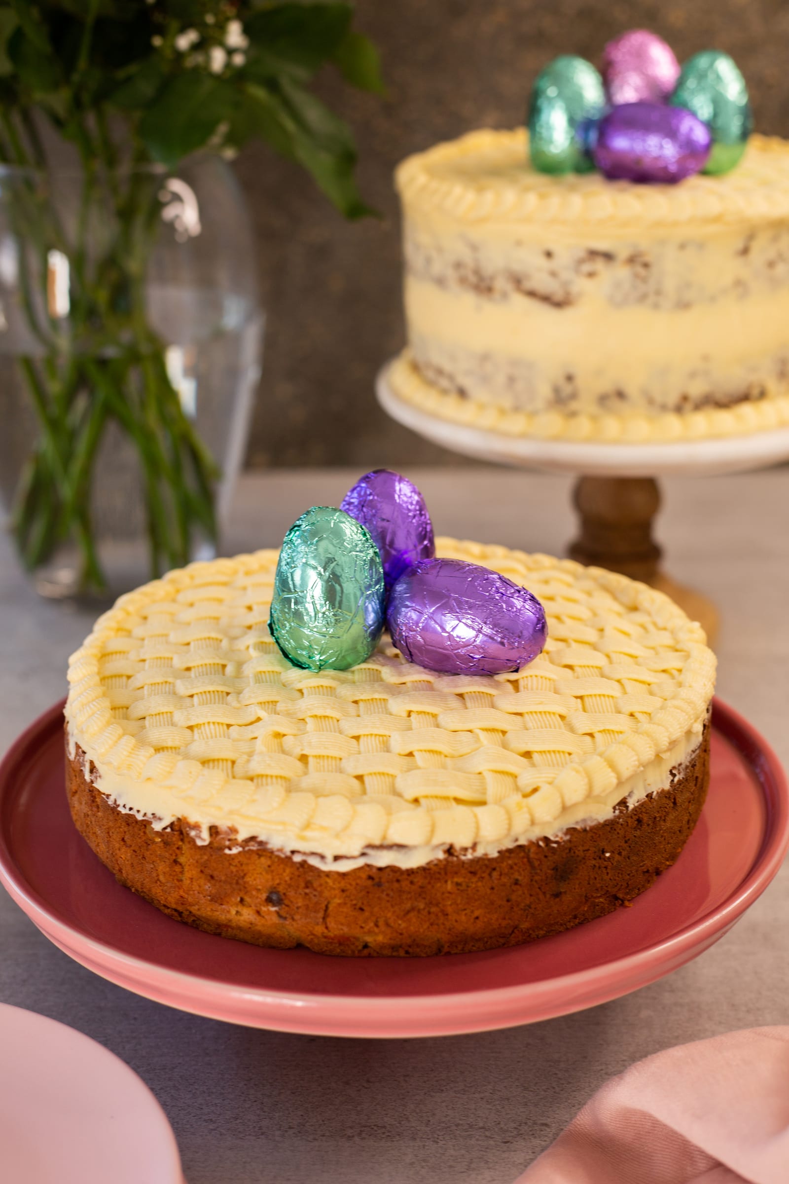 A gluten-free carrot cake with basketweave frosting on a pink stand, a layer cake behind
