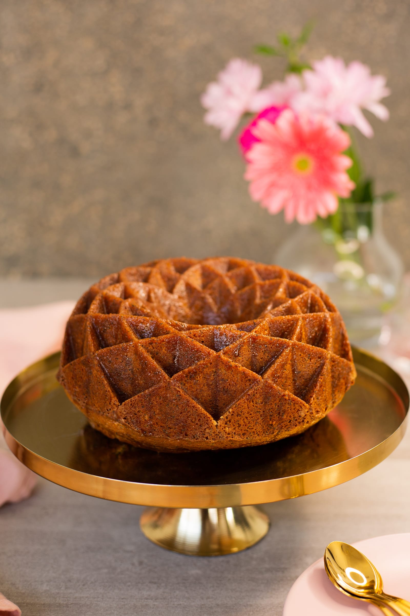 An unglazed lemon, poppyseed and olive oil bundt on a gold cake stand, pink flowers behind
