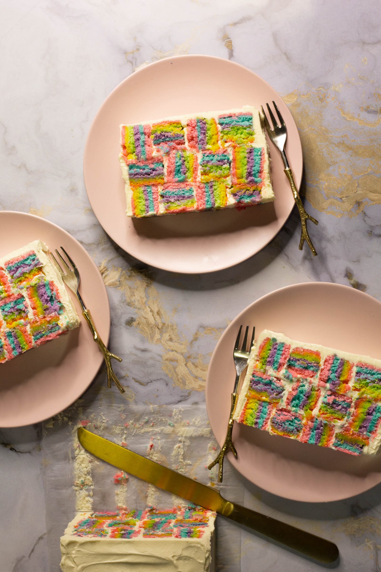 Slices of rainbow coconut layer cake on three pink plates with forks beside the cake, from above