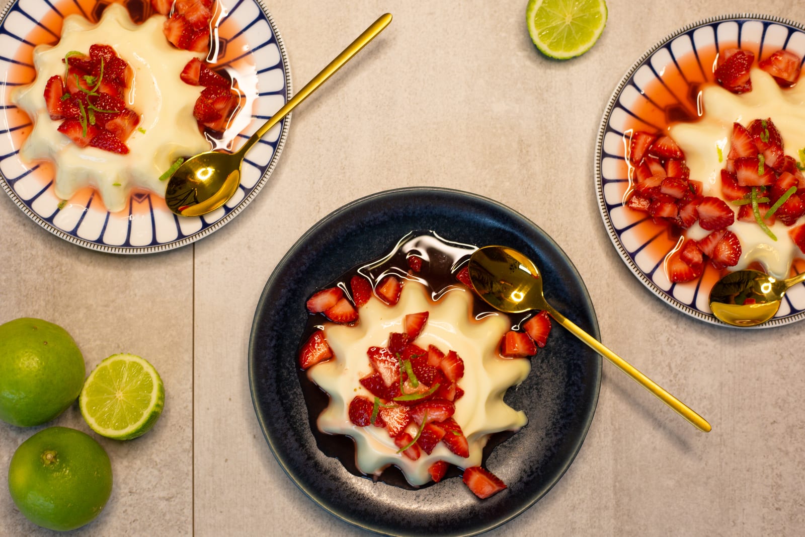 A lime panna cotta topped with strawberries on a dark blue plate, gold spoons and limes around it, from above