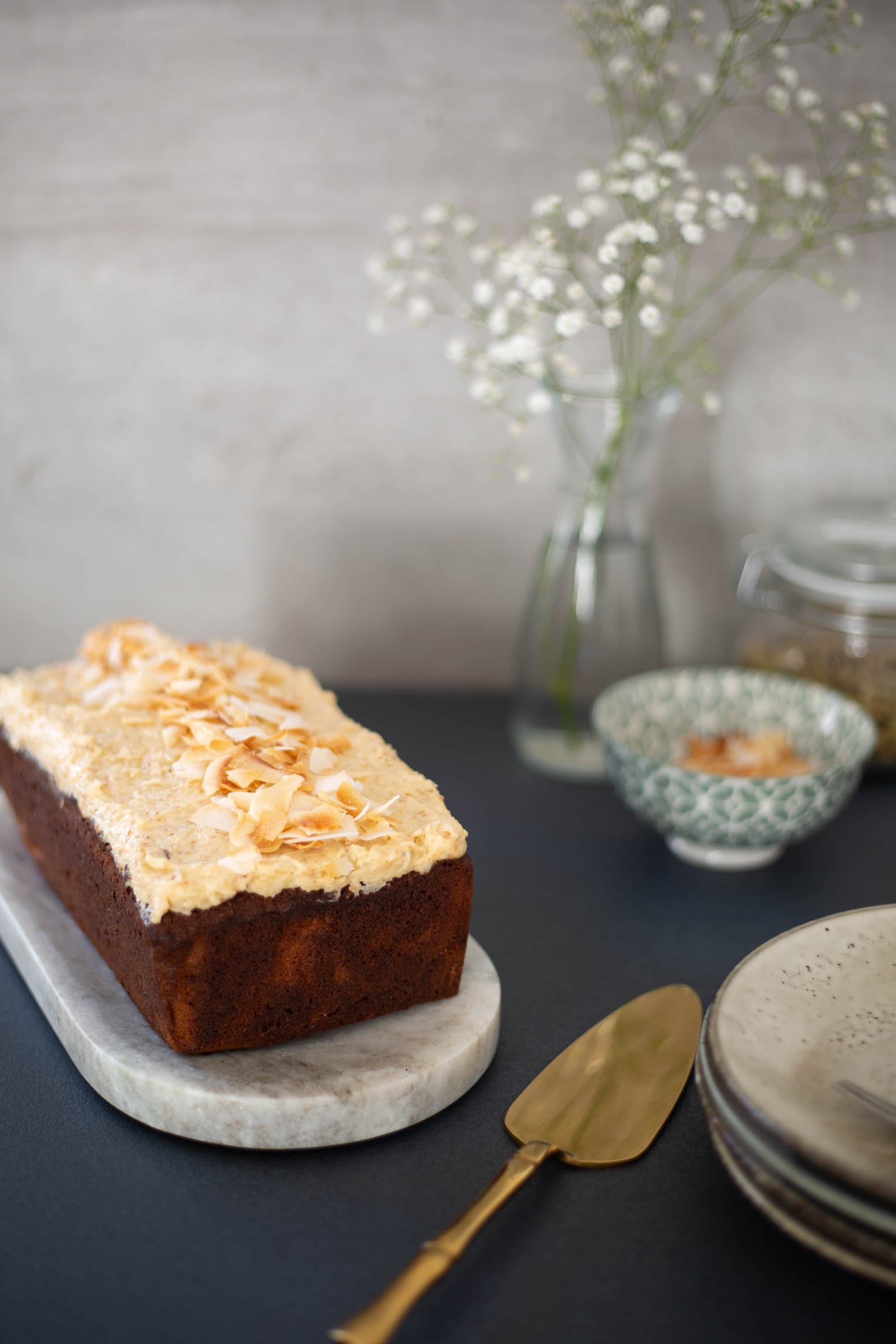 A whole gluten-free coconut loaf on a marble board, a vase of baby's breath behind