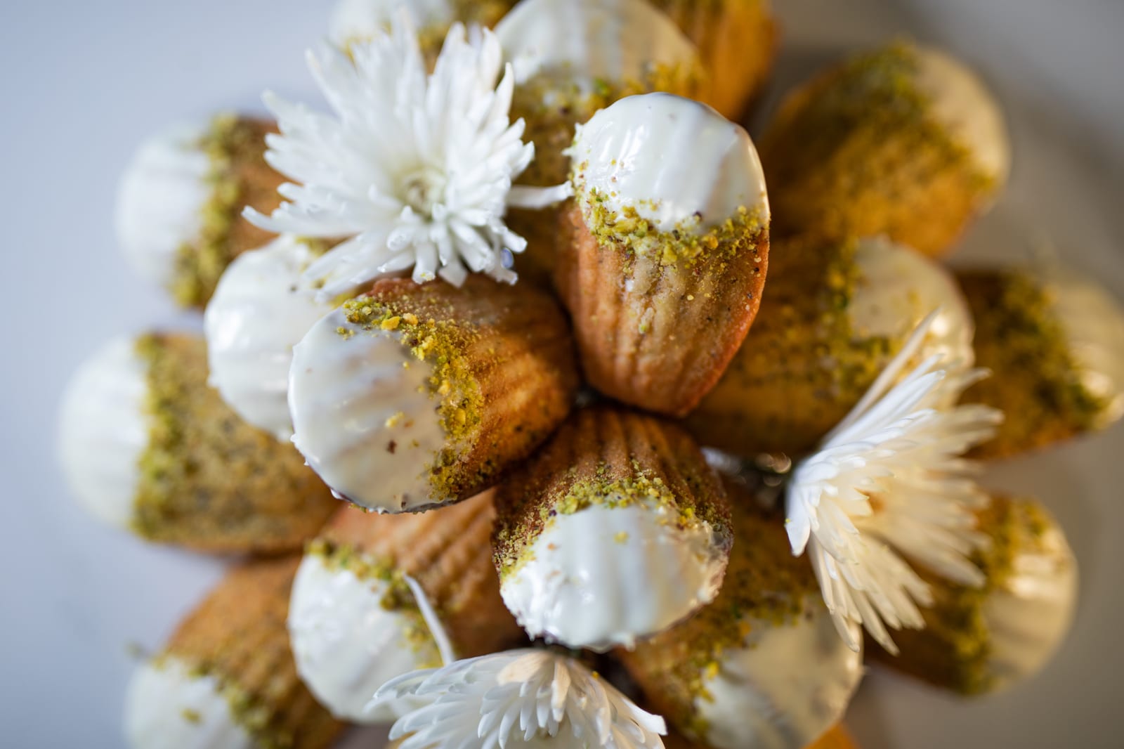 A mound of lemon and poppyseed madeleines dipped in white chocolate and pistachio, white flowers around, from above