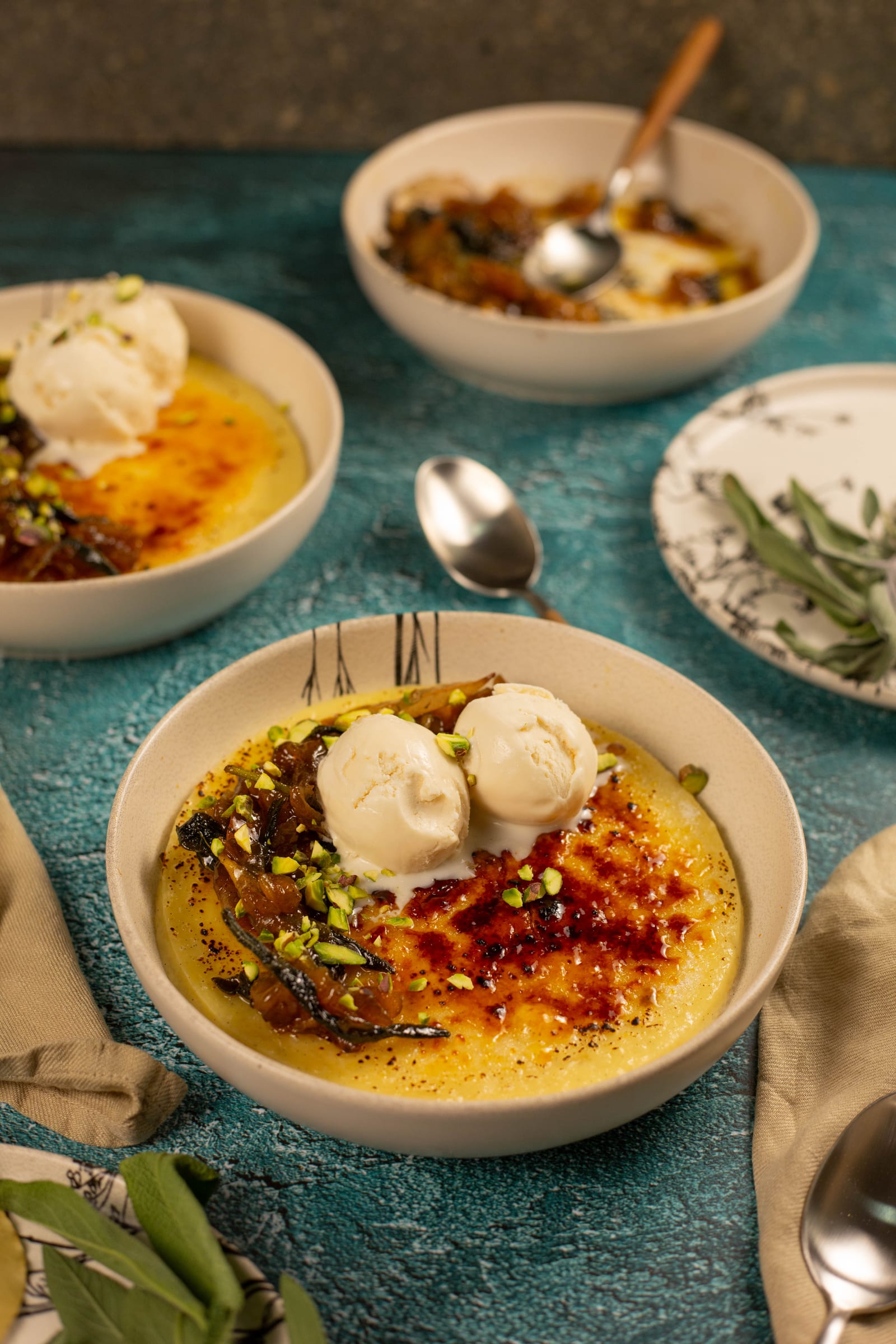 A bowl of rice pudding with ice cream and fried apples, with sage and spoons on a teal table
