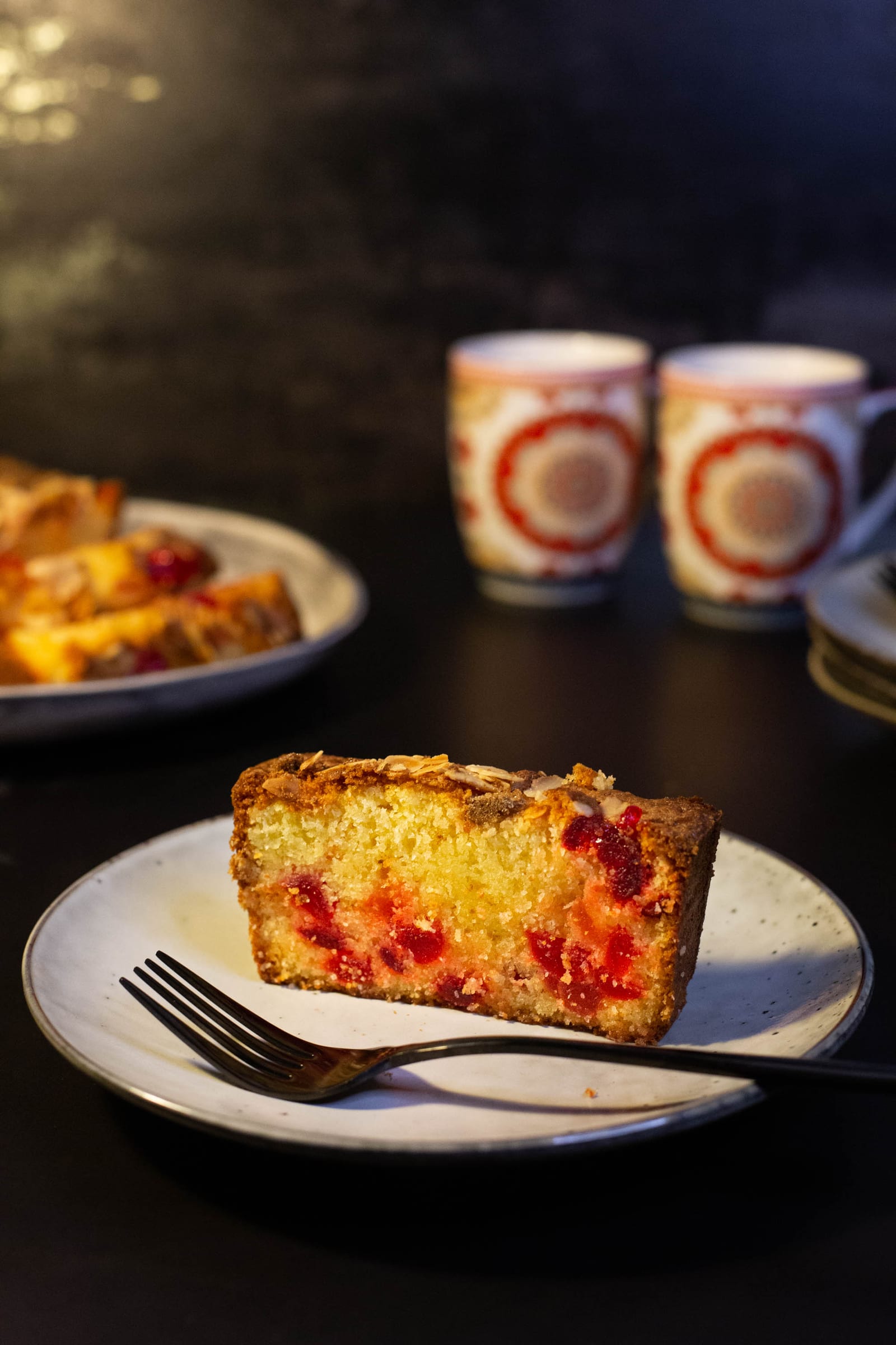 A slice of gluten-free cherry and almond loaf on a small plate with a fork, on a dark surface
