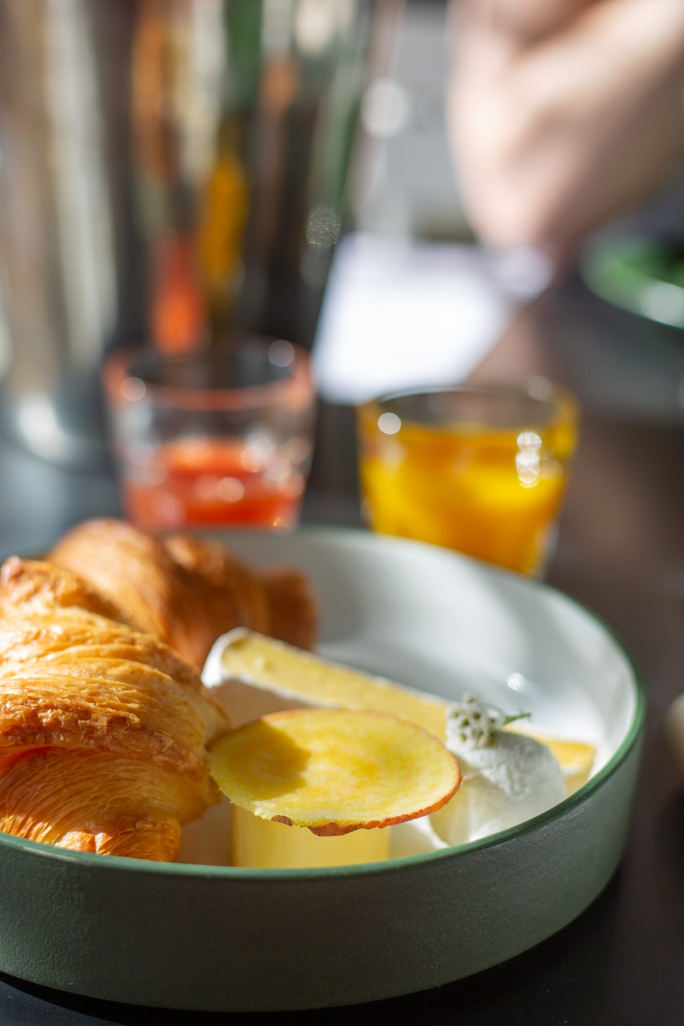 A croissant, brie and a golden beetroot slice in a green-rimmed bowl at Babel, with glasses of fresh juice behind