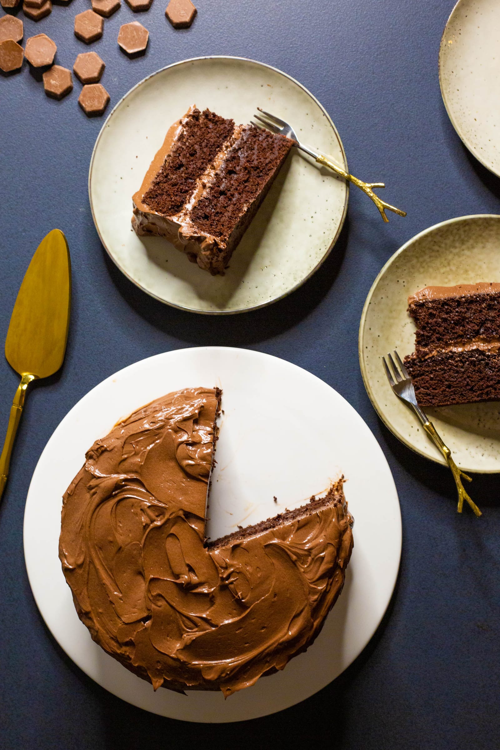 A cut chocolate fudge cake with slices on two small plates, a gold server and fork, from above
