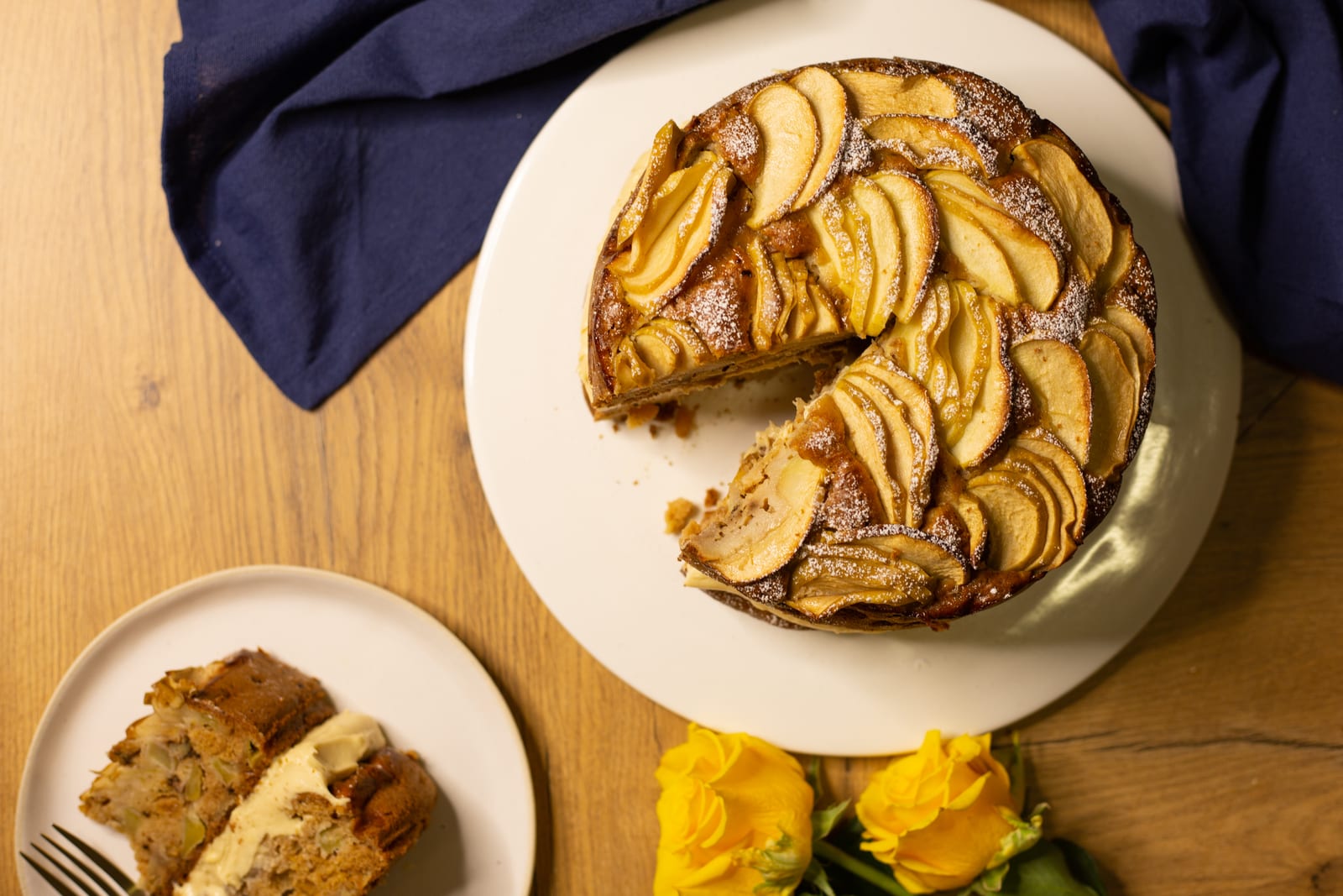 An apple and olive oil cake with a wedge cut out and a slice on a plate, yellow roses beside it, from above