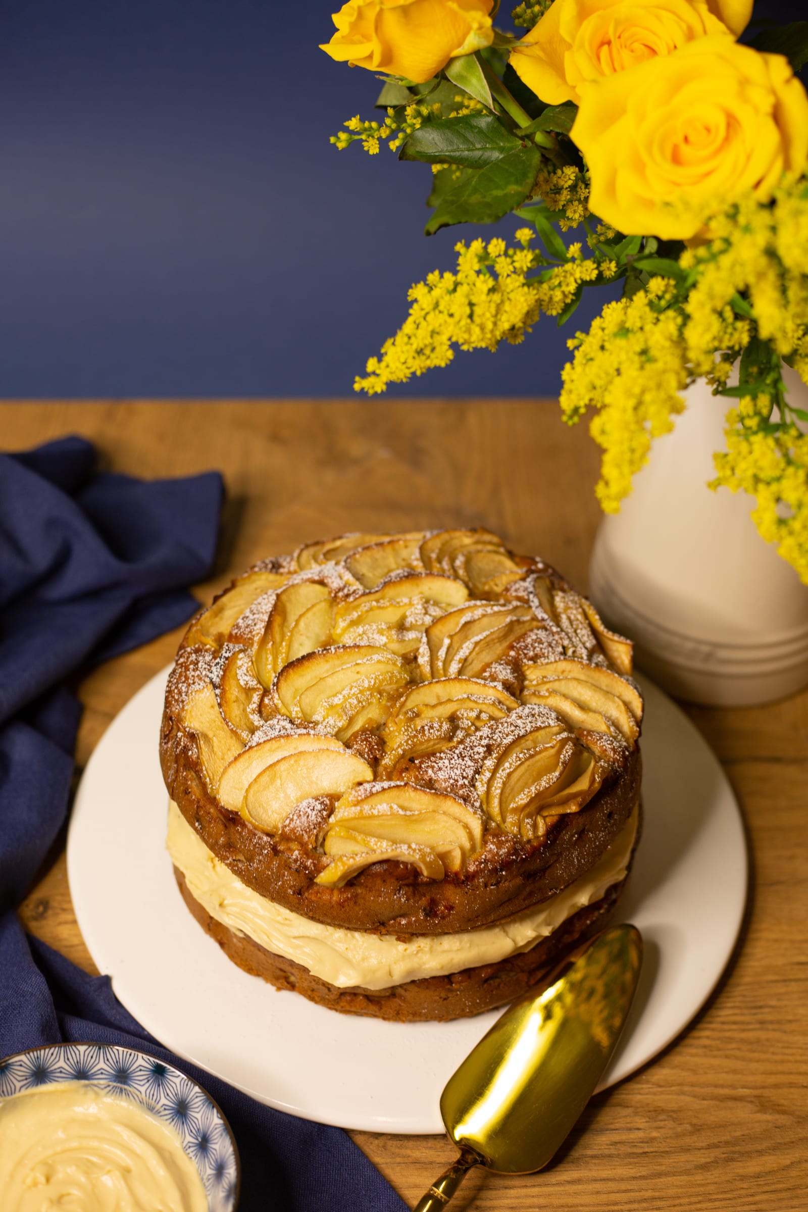 An apple and olive oil cake with a gold cake server and yellow flowers, from slightly above
