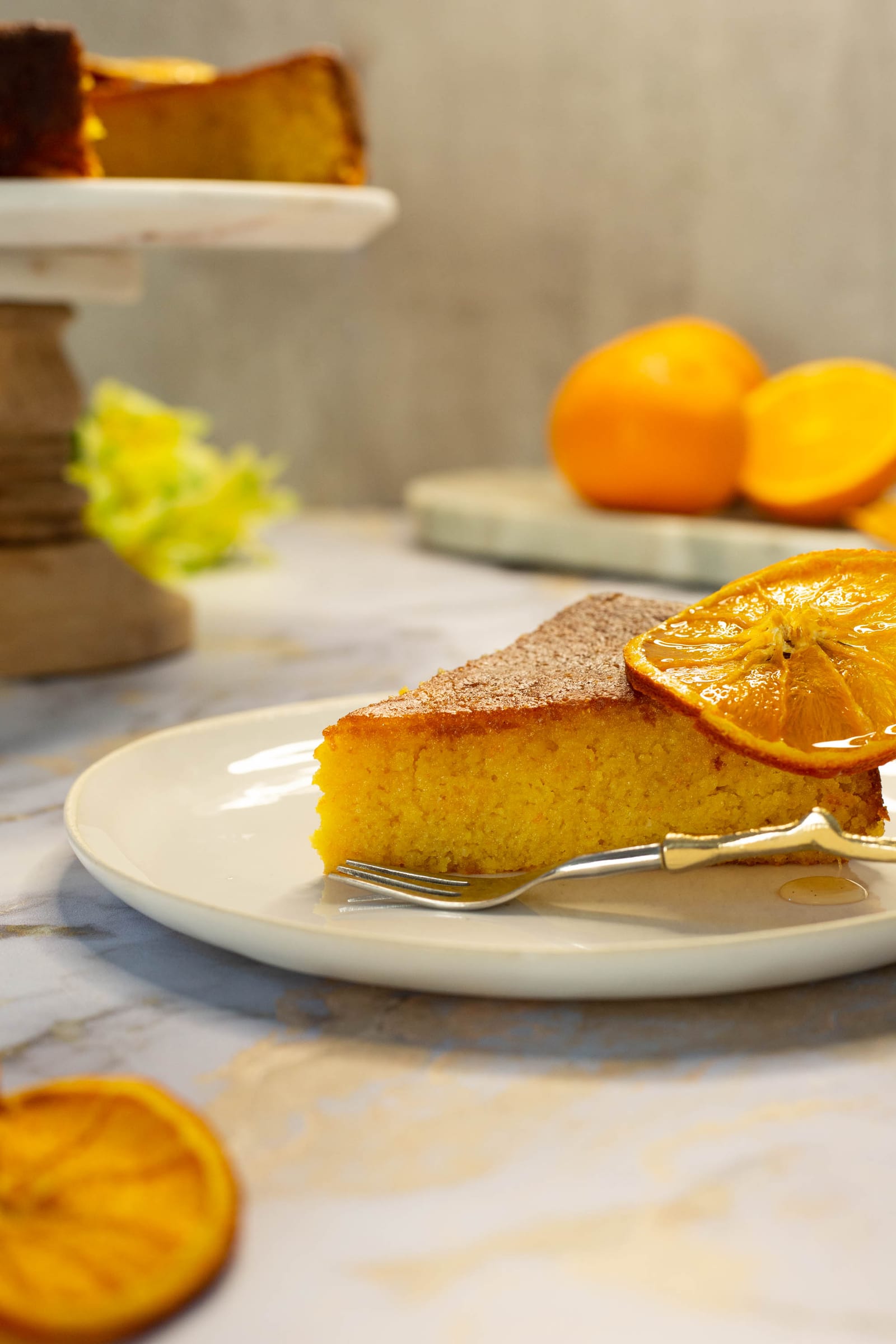 A slice of whole orange, cardamom and polenta cake with a candied orange on a white plate