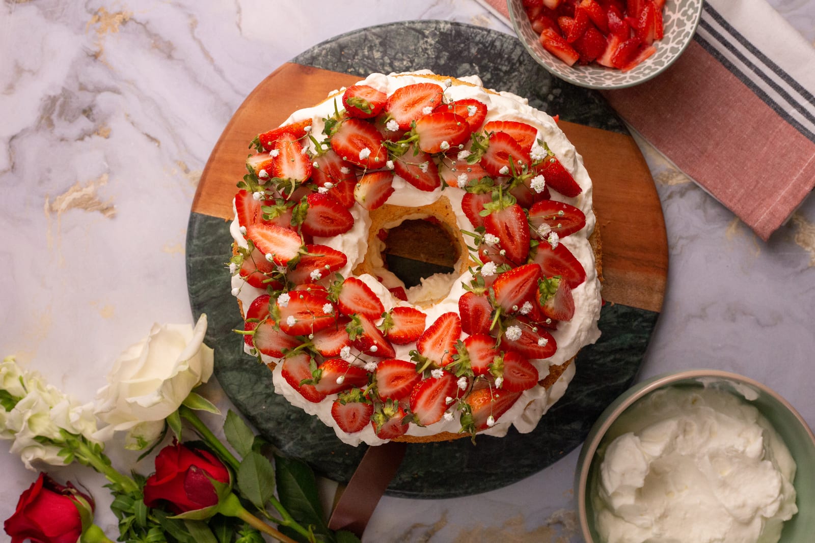 A strawberry and cream angel food cake topped with halved strawberries, roses and a pink striped cloth around it, from above