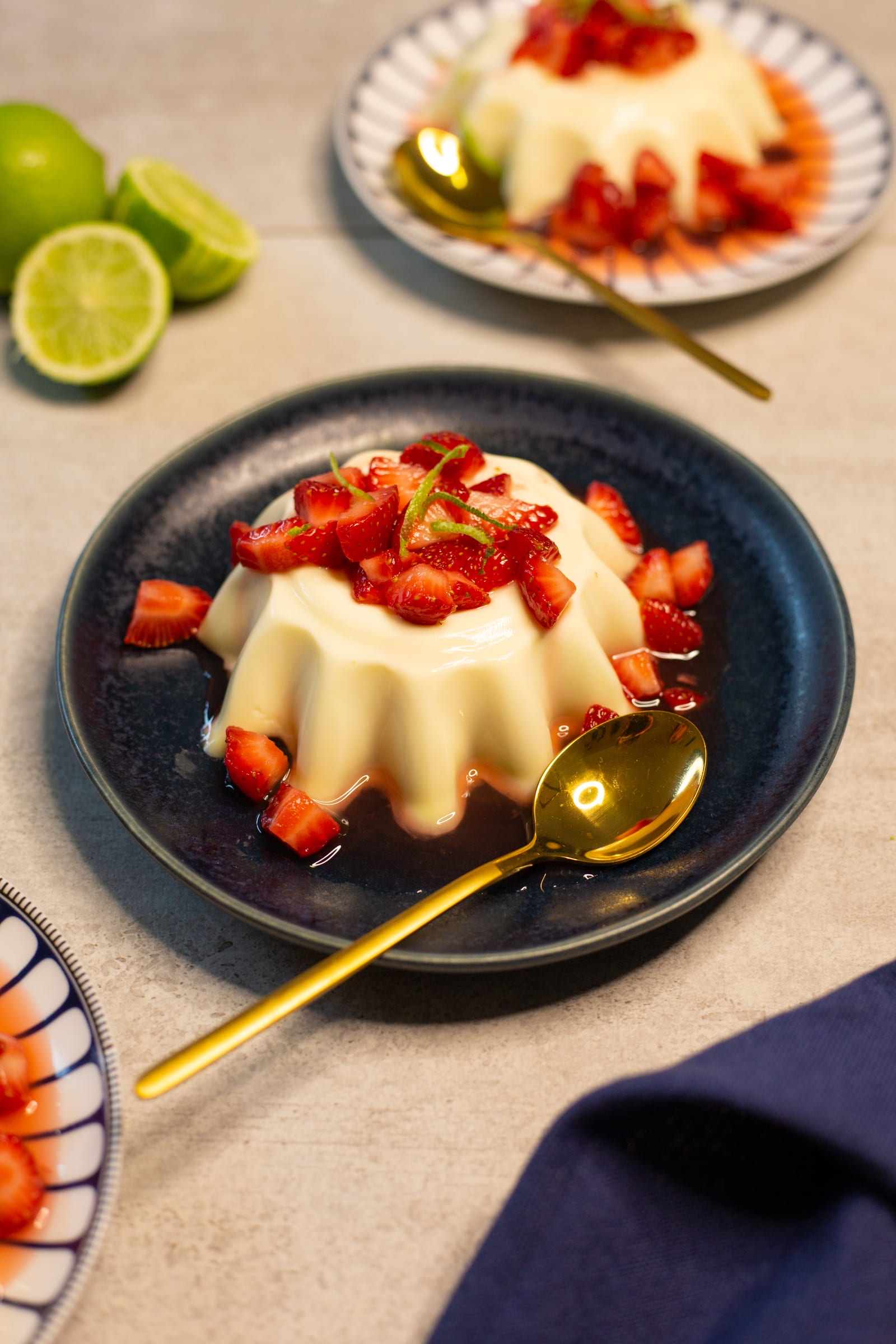 A lime panna cotta topped with strawberries beside a gold spoon and a navy cloth