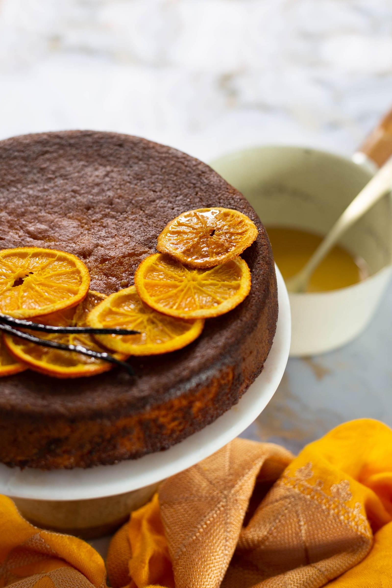 A whole orange, cardamom and polenta cake on its stand with a bowl of syrup and a spoon behind