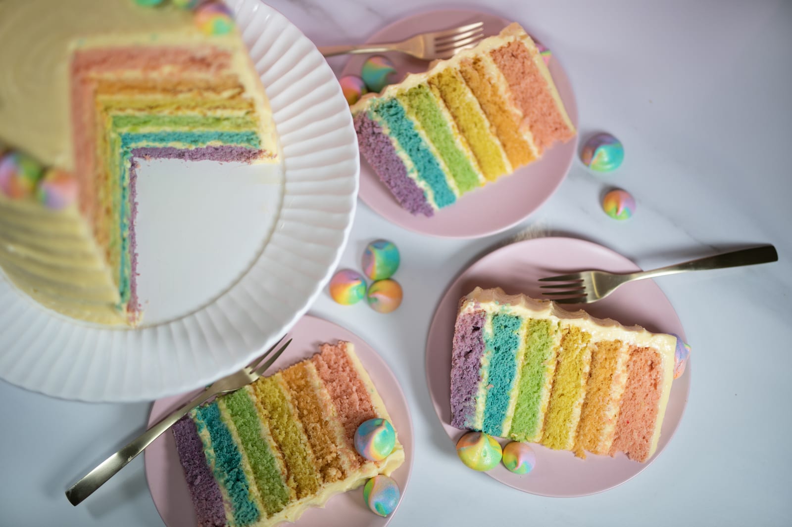 A cut pastel rainbow cake showing its rainbow layers, with slices on pink plates and forks, from above