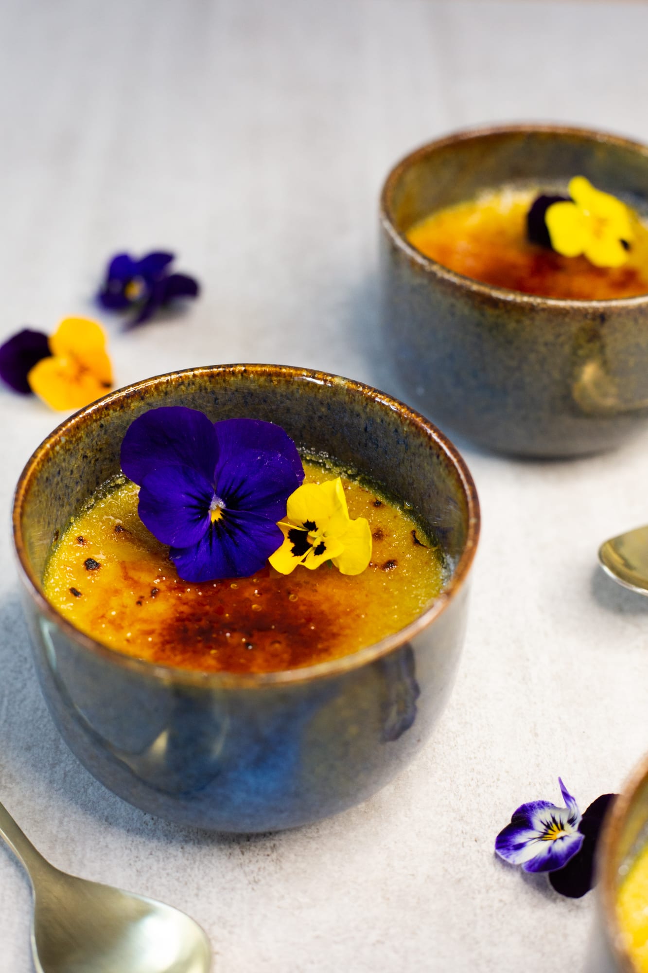A chai-spiced crème brûlée with a viola on its torched top, another ramekin behind